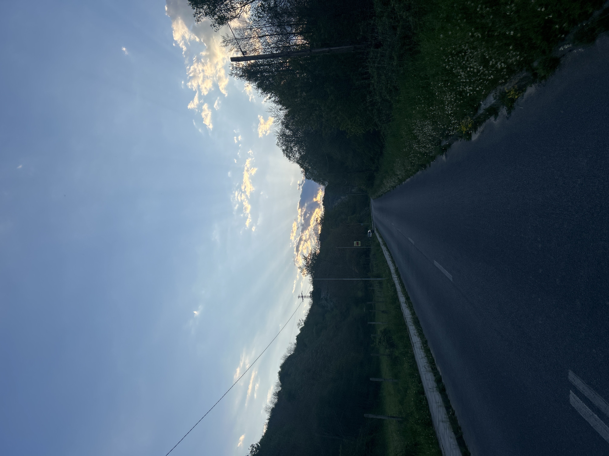 A roadside view in Zlatograd, Bulgaria is depicted. A paved asphalt road stretches into the distance, flanked on either side by green vegetation. On the left side, a wooded area climbs upwards, obscuring the view and creating a sense of enclosure. A utility pole and power lines are visible against the backdrop of the sky. The sky features a mix of blue and light, wispy clouds. Sunlight filters through the clouds, creating rays of light. The image has been rotated 90 degrees. The lighting suggests either early morning or late afternoon. The image appears to be taken from a moving vehicle.