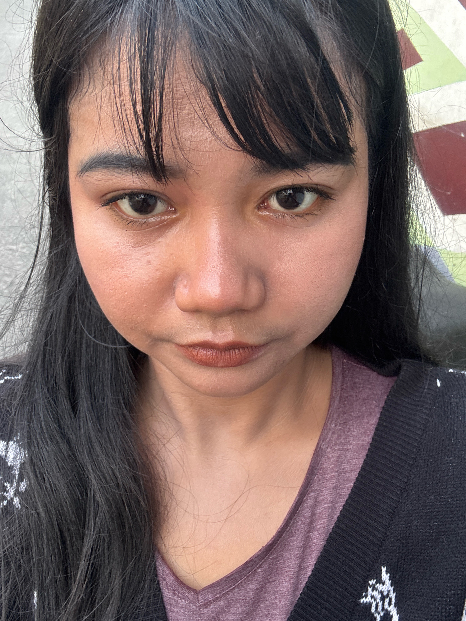 A close-up, head-and-shoulders portrait of a young woman with black hair and bangs is shown, facing the camera. Her skin appears slightly oily. She is wearing a maroon V-neck shirt under a black cardigan with white patterns. The background consists of a textured gray wall on the left and a colorful, geometric tiled wall on the right. The image was taken outdoors, likely in North Jakarta, Indonesia.