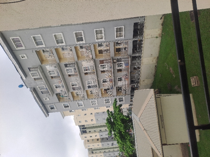 The image displays an urban residential landscape in Ikate, Nigeria, under an overcast sky. The central focus is a multi-story apartment building, approximately seven to eight visible floors tall, with a light gray exterior, white window frames, and white horizontal metal balcony railings. Interspersed balcony walls are painted light yellow. Multiple windows are visible, some open, some closed. Several balconies feature clotheslines with garments hanging. Air conditioning units are attached to the building's facade. A blue satellite dish is mounted on the highest visible roofline. To the left and in the background, additional similar multi-story residential buildings, primarily in gray and beige tones, extend into the distance. In the mid-ground, a single-story structure with a light beige wall and a light gray pitched metal roof is visible. This structure is bordered by a green grassy area and a light beige concrete retaining wall, which separates it from the base of the taller apartment building. A dense green tree stands to the left, partially obscuring some background structures. A dark metal railing occupies the immediate foreground, suggesting an elevated viewing position. No individuals are clearly discernible in active roles.