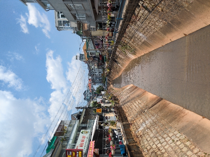 The image presents a concrete-lined canal, filled with murky brown water, cutting through an urban environment in Thành phố Đà Lạt, Vietnam. The canal extends straight into the distance, flanked on both sides by multi-story buildings, under a bright blue sky with scattered white clouds.
On the left bank, a row of buildings, primarily residential or mixed-use with ground-floor shops, features varied architecture, balconies, and numerous hanging flower planters. Overhead, a dense network of electrical wires is visible. A prominent white sign with green text reads "ĐẬU ĐÀ LẠT," and a partial sign indicates "PHAT." Awnings are extended over some shopfronts.
The right bank exhibits similar multi-story buildings, some with modern glass enclosures and balconies displaying drying laundry. A blue metal railing runs along the canal's edge, also adorned with hanging flower planters. Electrical wires are also present overhead on this side.
The canal walls are constructed with a lower concrete section and an upper layer of rough-hewn stone blocks, with sparse vegetation. In the background, more buildings ascend a hillside, and a notable structure with a peaked roof and a solar water heater is visible centrally, further down the canal's path where it passes under a distant bridge. No individuals, direct actions, or interactions are observable within the scene, which is illuminated by direct sunlight.