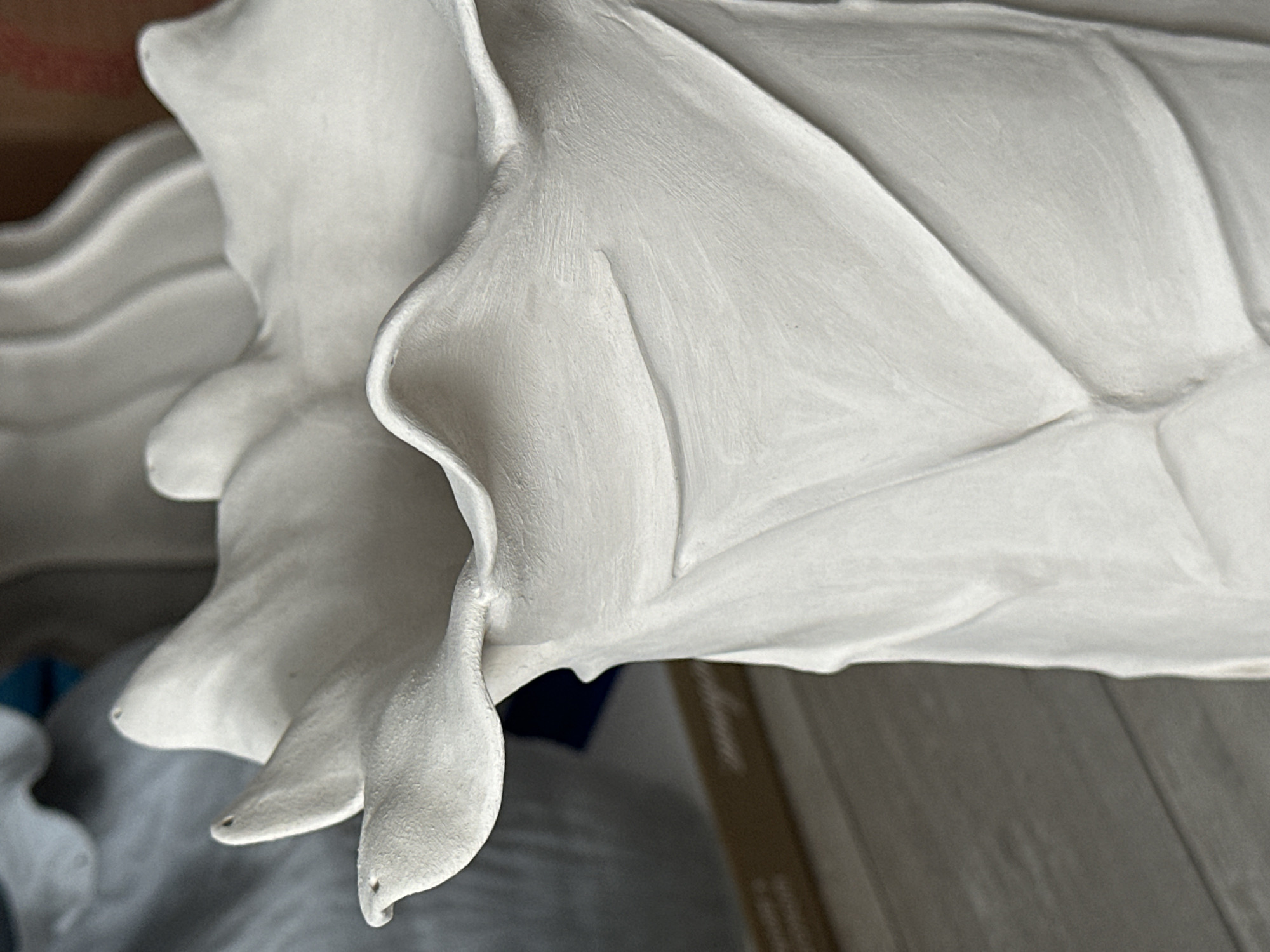 The image shows a close-up of a white, unfinished ceramic vase with a ruffled top and textured, petal-like detailing on its body. The vase has a matte finish. In the background, out of focus, are other similar ceramic pieces and a blurry wooden surface. There is no indication of the city of Nevinnomyssk, Russia, or any people, actions, or interactions.