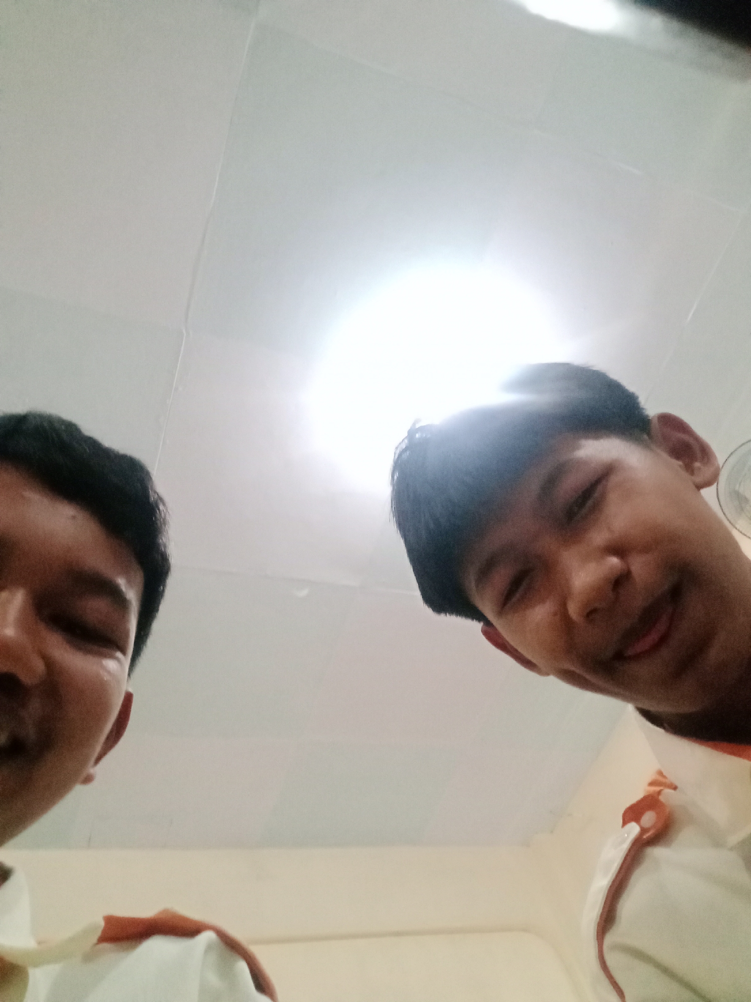 Two young males are captured in a low-angle selfie taken indoors, possibly in a classroom or common area in Secang, Indonesia. The ceiling is visible, featuring a bright light fixture and a tiled pattern. The male on the left is partially in frame, with his face turned towards the camera. The male on the right is more fully visible, looking directly at the camera with a slight smile and his tongue peeking out. Both are wearing white shirts with orange trim on the collar and shoulder area, suggesting a school uniform or team attire. The overall lighting is bright, with some lens flare from the overhead light.