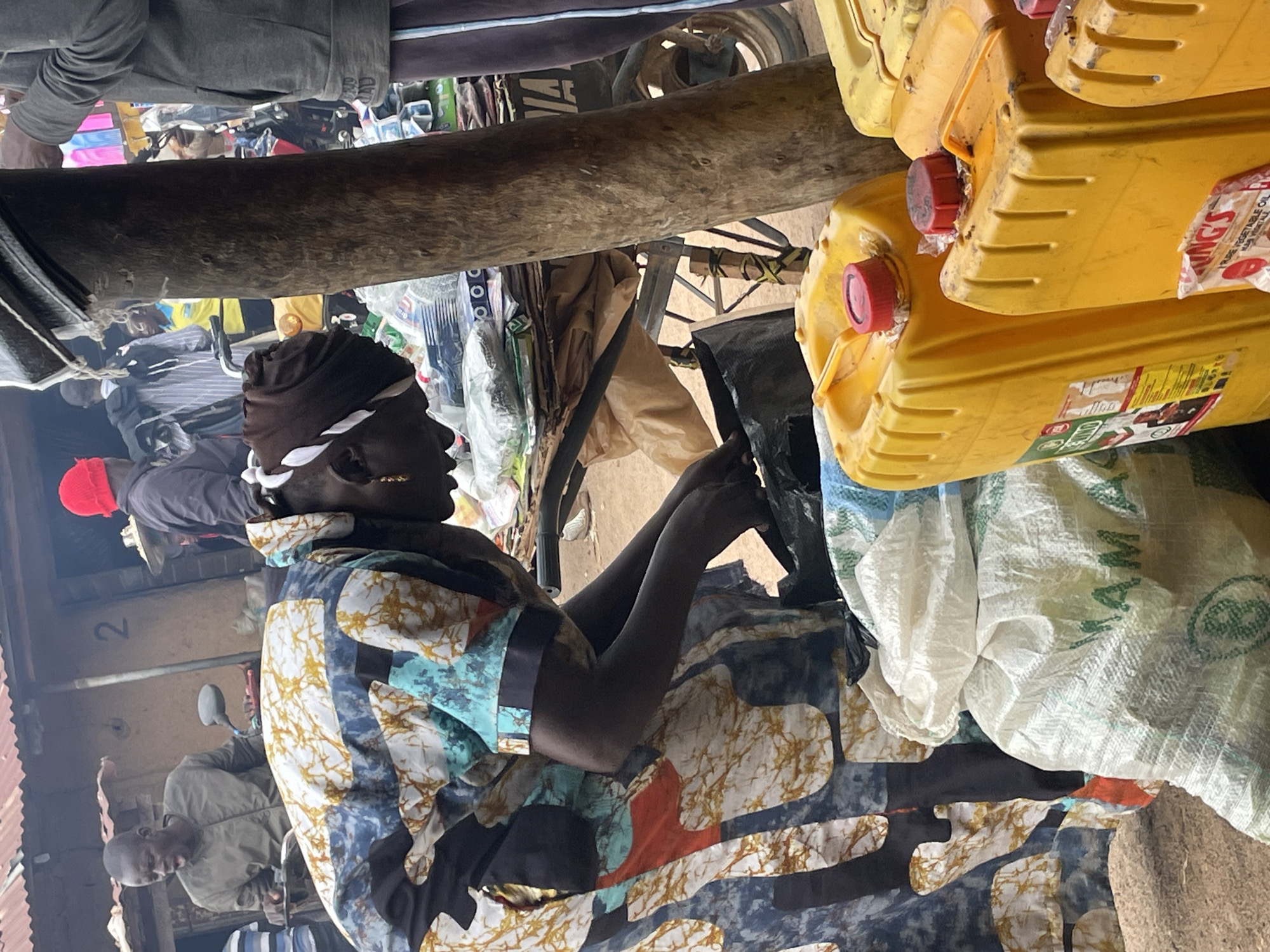The image depicts a bustling marketplace scene in Wang, Nigeria. In the foreground, a person wearing a colorful patterned dress with a headscarf is interacting with a cart laden with goods. They are reaching into a black bag. Behind them, several large yellow plastic containers, likely for oil or other liquids, are stacked. A white sack with text, possibly for agricultural products, is also visible. The background reveals more market stalls, people, and a building with the number '2' visible. The overall impression is one of active commerce in a lively urban setting.