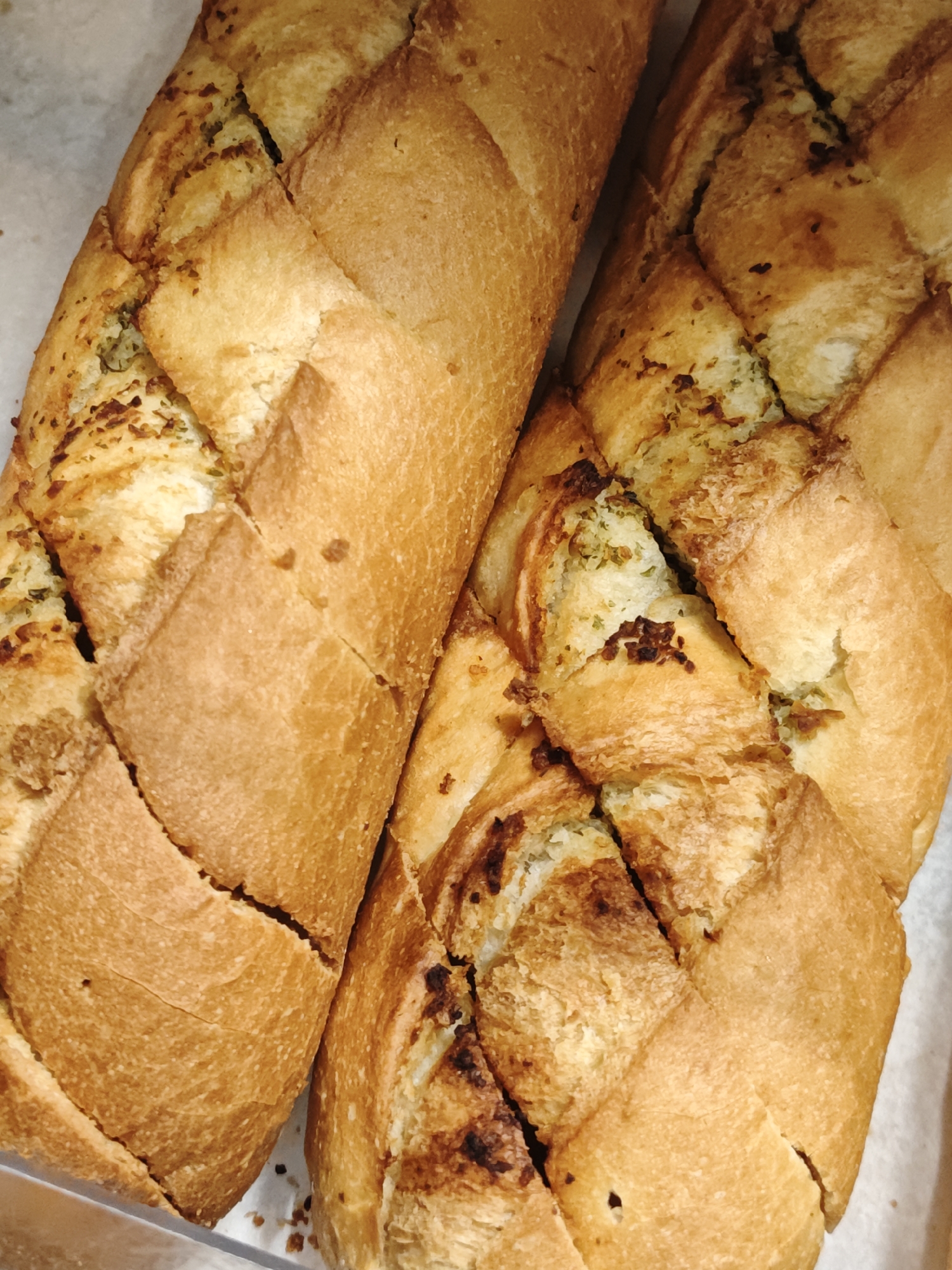 The image shows a close-up view of two loaves of freshly baked bread, likely garlic bread, with visible herbs and seasoning baked into the crust. The bread has a golden-brown, crispy exterior with diamond-shaped scores on top, revealing the fluffy interior. The lighting suggests an indoor setting, possibly a bakery or kitchen. There are no people or any indication of the city of Shcherbinka, Russia present in the image.
