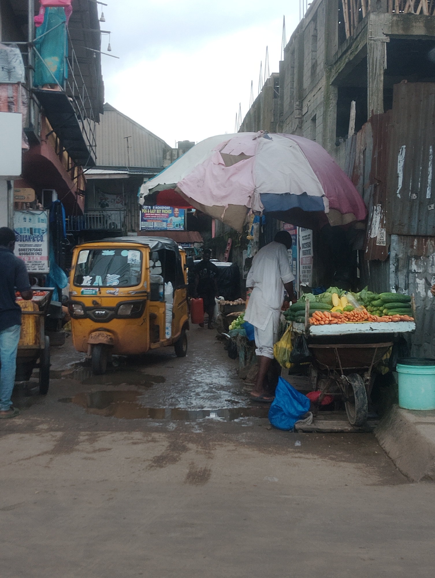 This image captures a street scene in Iju, Nigeria, likely a market area. A yellow auto-rickshaw, a common mode of transport in Nigeria, is parked on the wet, uneven pavement. In the foreground, a person is standing next to a cart filled with produce, including cucumbers, carrots, and corn. Another person, wearing a white traditional outfit, is attending to the produce stand. Buildings line the street, some appearing unfinished with exposed rebar and corrugated metal siding. Overhead, tarps and umbrellas provide shade, and various signs, some with text that is difficult to read, are visible on the buildings. The overall atmosphere suggests an active, outdoor marketplace with a slightly overcast sky.
