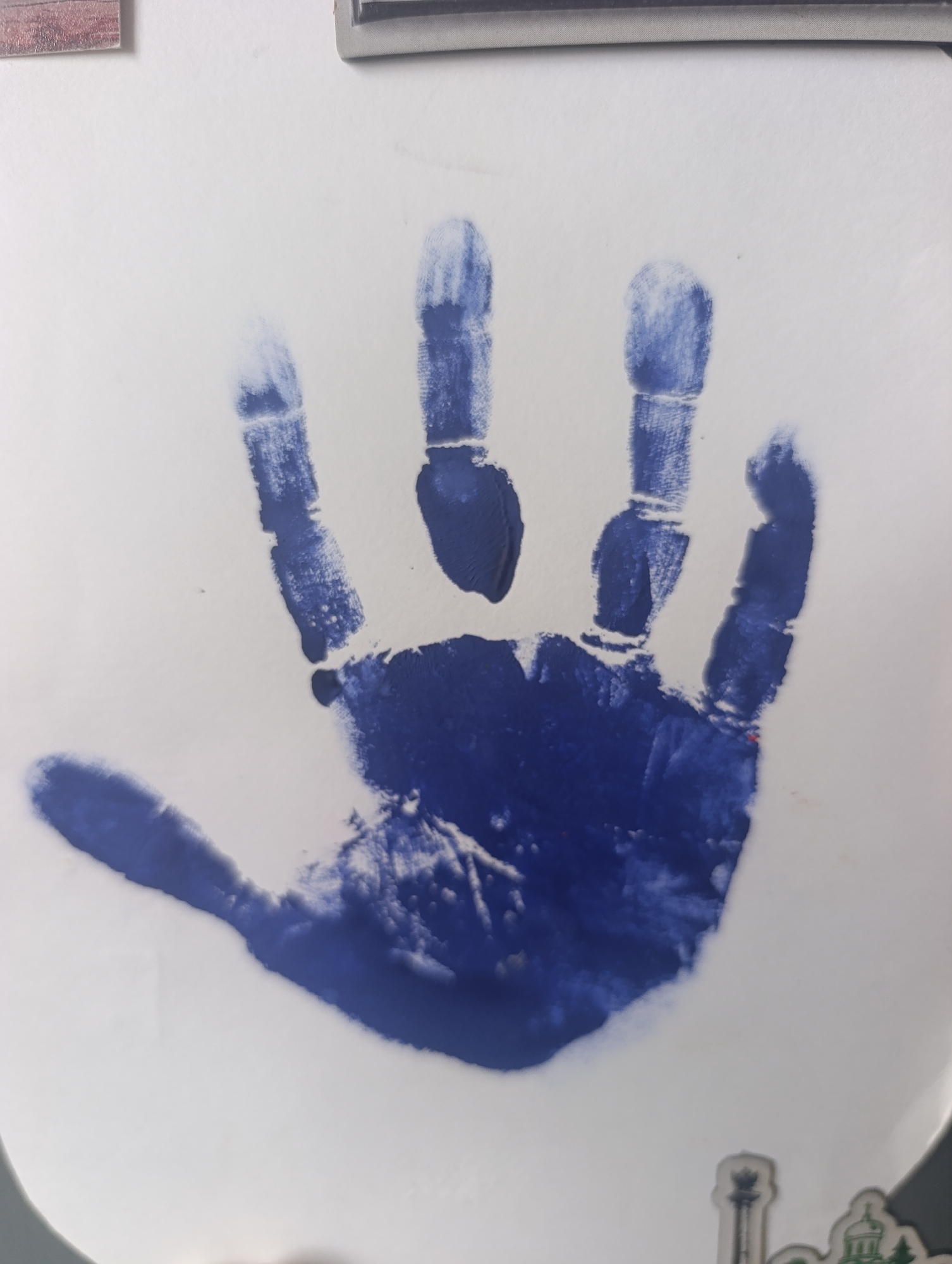 A blue handprint is centered on a white surface. The handprint is clearly visible, with the palm and five fingers distinctly outlined in blue paint. The texture of the paint suggests it was applied with some pressure, leaving slight variations in thickness and coverage. The background is plain white, with no discernible details or context. The image does not provide any information related to Naro-Fominsk, Russia, or any specific location within a city. There are no people, actions, or interactions depicted in the image. The focus is solely on the handprint itself.