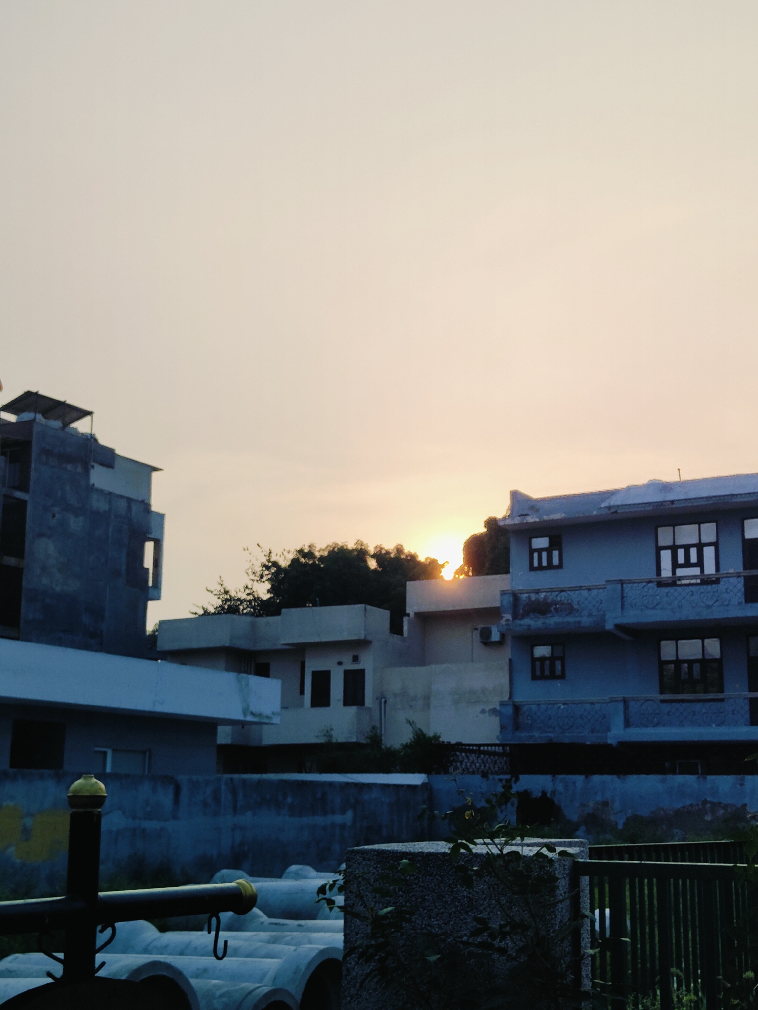 This is a full-shot, eye-level, outdoor, vertical photograph taken at dusk in Gurgaon, India. The sky is a pale orange-yellow, indicating the sun is low on the horizon. In the mid-ground, several buildings are visible. On the left, there is a multi-story building with unfinished construction, grey concrete walls, and visible rebar. To the right of this, a lighter colored building with dark-framed windows is partially obscured by trees. Further to the right, a blue multi-story building with balconies and dark-framed windows stands out. In the foreground, a dark metal structure with a yellow circular top is visible, along with a stack of large concrete pipes. The overall scene depicts an urban residential or construction area at sunset.