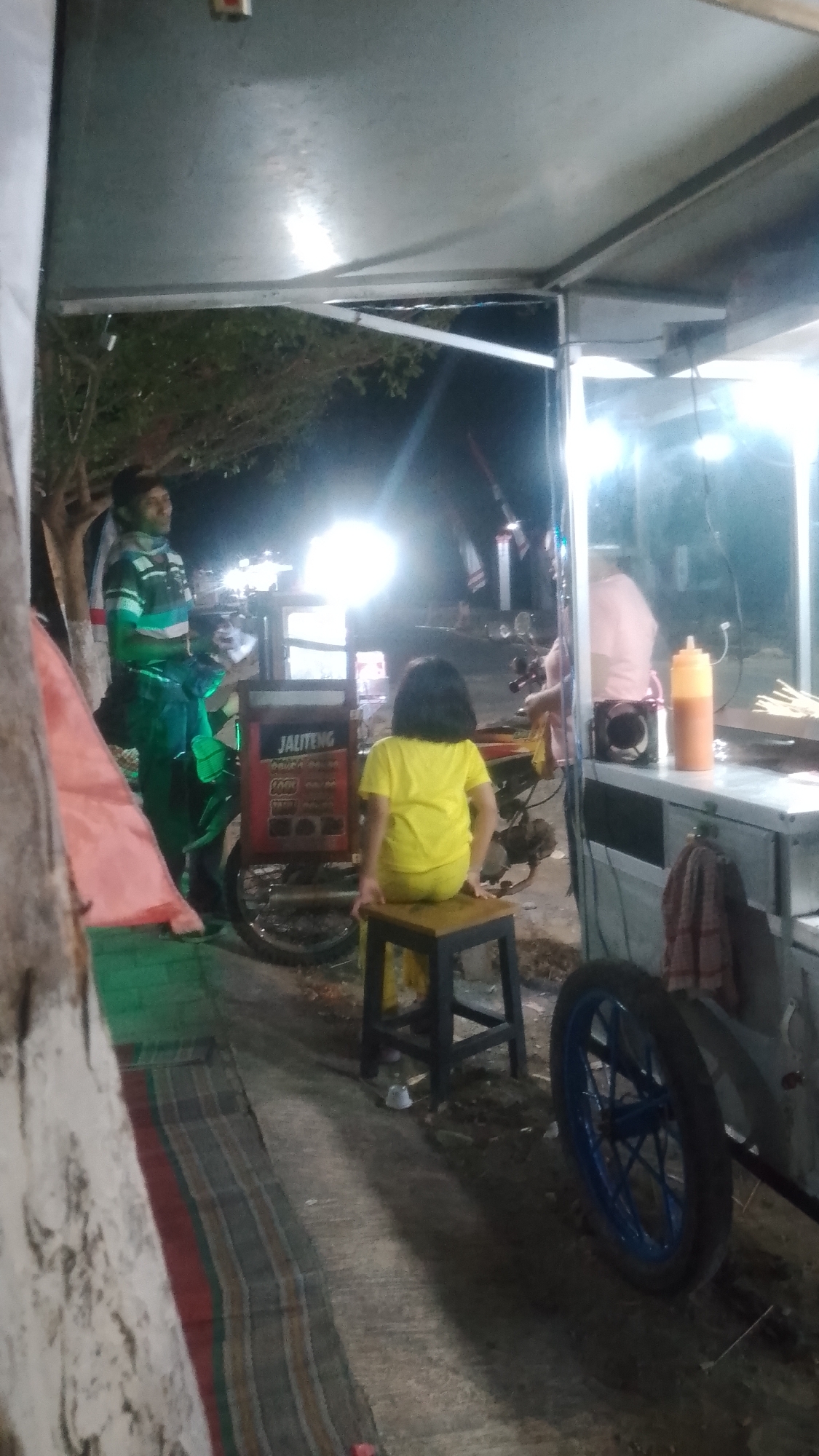 In the city of Karanglangu, Indonesia, a night scene unfolds around a food cart. A young girl in a bright yellow shirt and pants sits on a small stool, facing away from the camera, her back towards a food cart. The cart, adorned with a sign that reads "JALITENG," is illuminated by a bright light. To the left of the cart, a man wearing a striped shirt and dark pants stands beside a motorcycle, which appears to be integrated with the cart. A colorful woven mat is spread on the ground in the foreground. In the background, another person in a light pink shirt can be seen behind the food cart. The scene is lit by artificial lights, creating a warm glow and casting shadows on the ground.