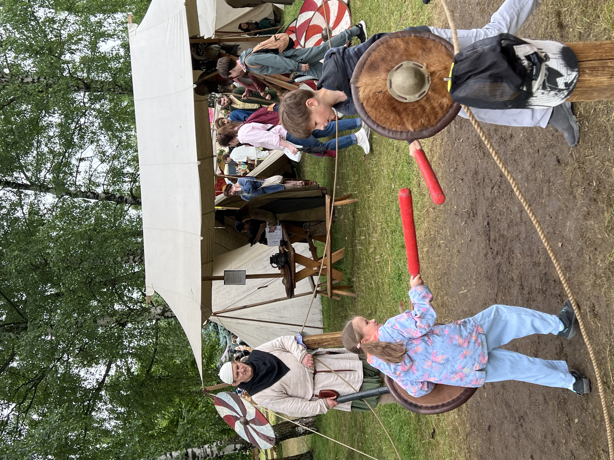 This image depicts an outdoor historical reenactment event. In the foreground, two children are engaged in a mock sword fight, each holding a red foam sword and a round shield. The girl on the left wears a blue floral jacket and light blue pants, while the boy on the right is in a grey hooded jacket, light grey pants, and dark grey shoes. Behind them, an adult male dressed in historical attire, including a white cap, black scarf, and light beige tunic over a green skirt, stands near a wooden post. In the background, several tents are set up, suggesting a festival or fair. People in various historical costumes are visible milling around the tents and a wooden table. The ground appears to be muddy grass. The overall scene suggests a family-friendly event with a focus on history. No specific landmarks or indications of Moscow, Russia are present in the image.