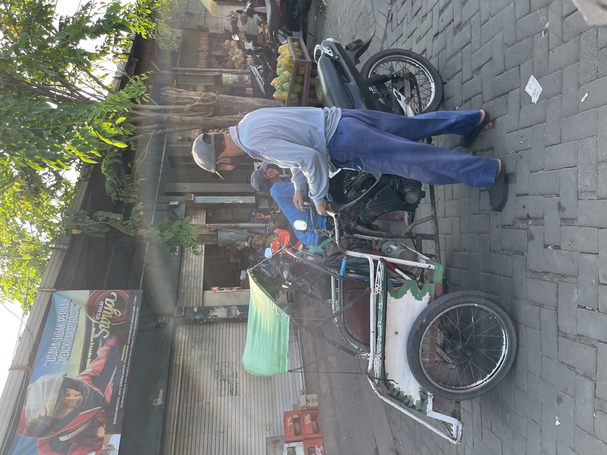 The image depicts a street scene in Juwet Kenongo, Indonesia. In the foreground, a man wearing a grey cap, a light blue long-sleeved shirt, and blue pants stands next to a three-wheeled vehicle that appears to be a modified motorcycle with a passenger carriage. The vehicle has a green canopy and white and green detailing. The man is facing away from the camera, looking towards the passenger carriage. He appears to be interacting with the vehicle or preparing to operate it. In the background, there are several other motorcycles, a fruit stand displaying papayas, and shops with closed roll-up doors. A banner is visible hanging above the shops with text in Indonesian, featuring an image of a person in a racing helmet. Trees with green foliage are also present, casting shadows on the scene, indicating it is likely daytime. The ground is paved with brick-like stones.