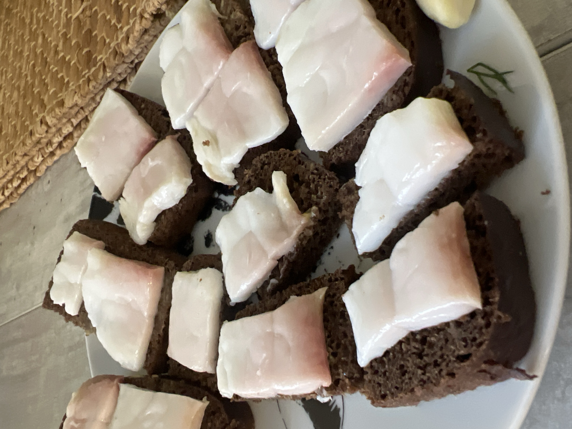 This is a close-up, eye-level, indoor shot of a plate of black bread slices topped with pieces of cured pork fat. The bread is dark brown and appears to be pumpernickel or a similar rye bread. The pork fat is white with pinkish edges, suggesting it is thinly sliced salo. The pieces of fat vary in size and shape, with some being rectangular and others more irregular. The plate has a black and white abstract design around the rim. In the background, to the right, a woven placemat with a coarse texture is visible. The lighting is natural and comes from above, casting soft shadows. The overall impression is that of a rustic, traditional dish. The image does not provide any clues or information that would allow for determining the location in the city of Volgograd, Russia.