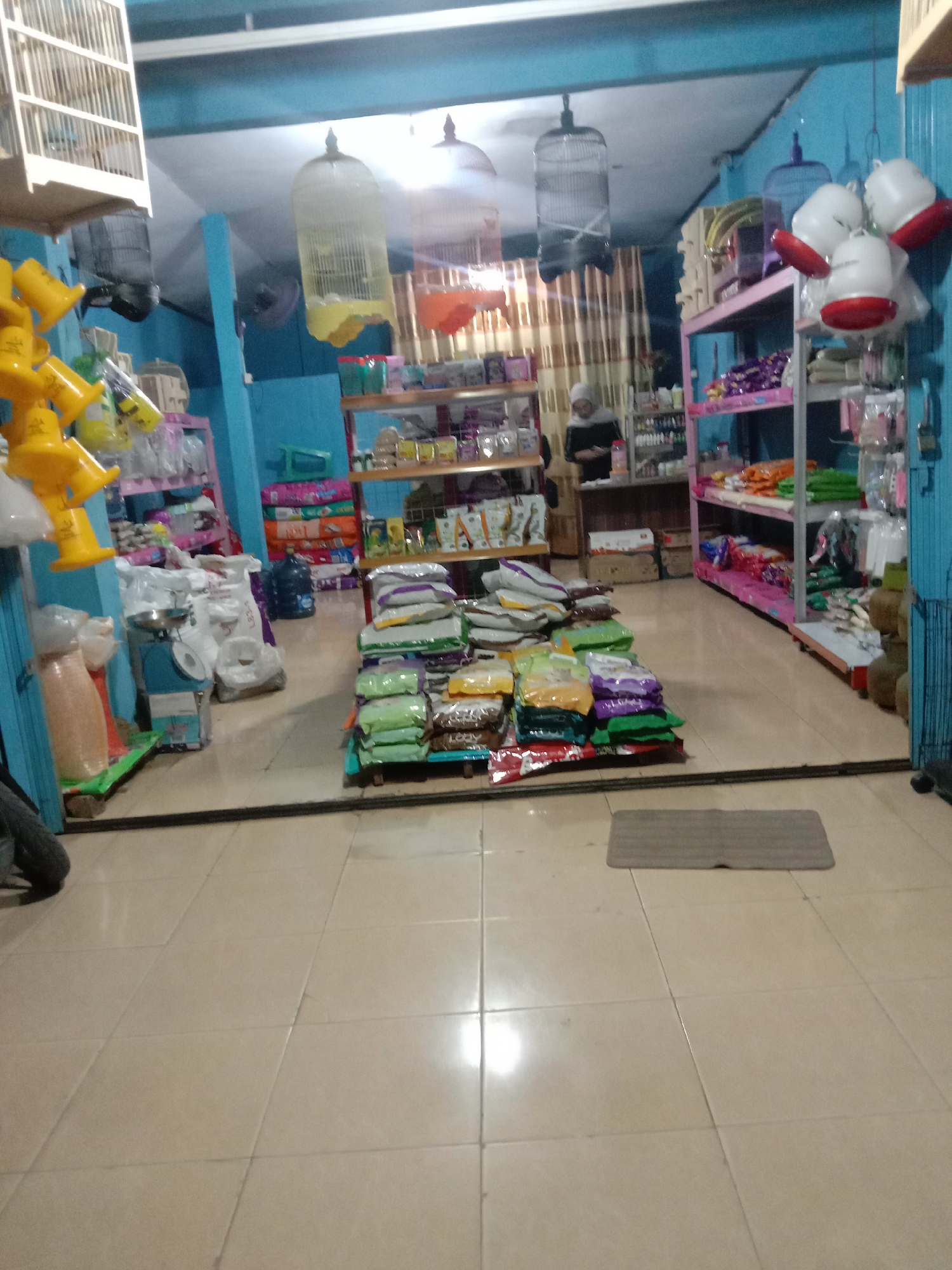 The image depicts the interior of a small retail store, likely a pet supply shop, located in Fajar Mataram, Indonesia. The store is filled with shelves stocked with various products, including bags of pet food, bird cages, and other pet-related items. In the background, a person wearing a headscarf and dark clothing appears to be attending to customers or organizing merchandise. The flooring is tiled, and the walls are painted a light blue. The lighting suggests it might be evening or an indoor environment with artificial illumination. The overall impression is one of a bustling local business.