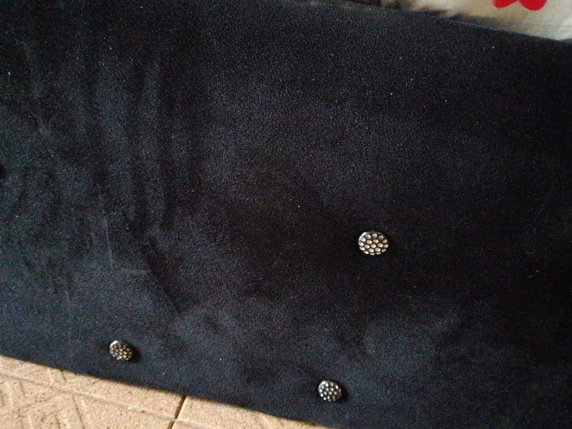 A close-up, top-down, eye-level shot shows a dark, textured fabric, possibly velvet or a similar material. The fabric is adorned with four small, circular embellishments, each appearing to be a cluster of tiny, reflective beads or rhinestones. Three of these embellishments are visible near the bottom of the frame, with the fourth located in the upper right quadrant. The lighting casts a slight sheen on the fabric and highlights the reflective surfaces of the embellishments. The background appears to be a tiled surface, possibly a floor, in a neutral tone. The scene is indoors, in an unknown location.