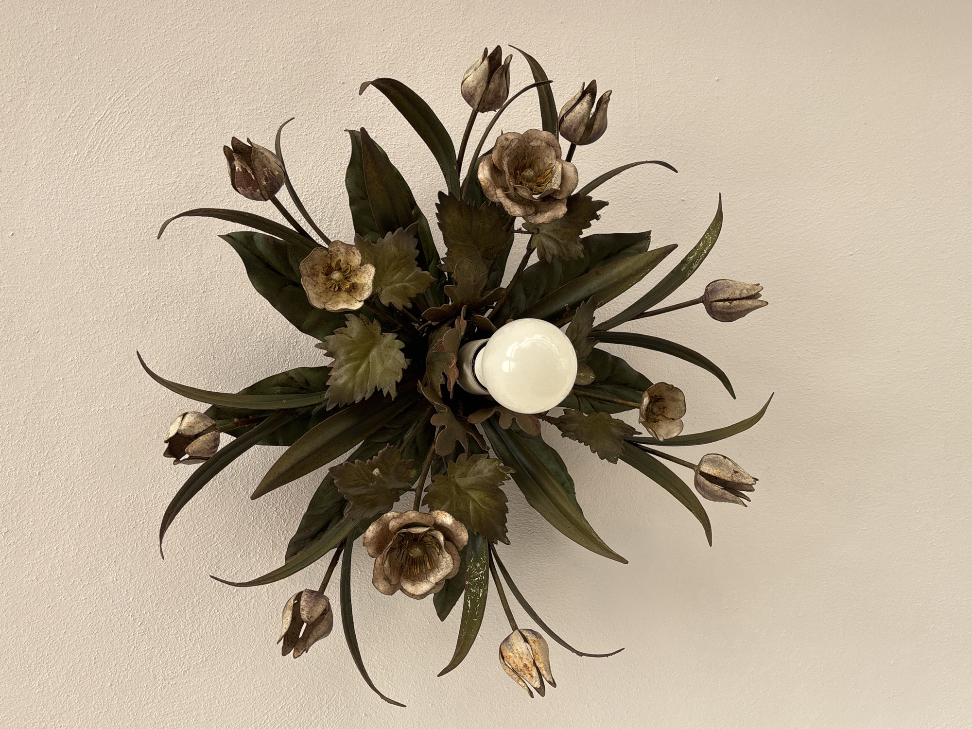 A ceiling light fixture, designed to resemble a floral arrangement of metal tulips and leaves, is attached to a textured, off-white ceiling. The fixture has a central, exposed white LED light bulb. The metal flowers and leaves are a dark green and metallic bronze color, with some showing a distressed or aged finish. The arrangement is symmetrical and radiates outwards from the central bulb. The image was taken in Orihuela, Spain, in an indoor residential setting.