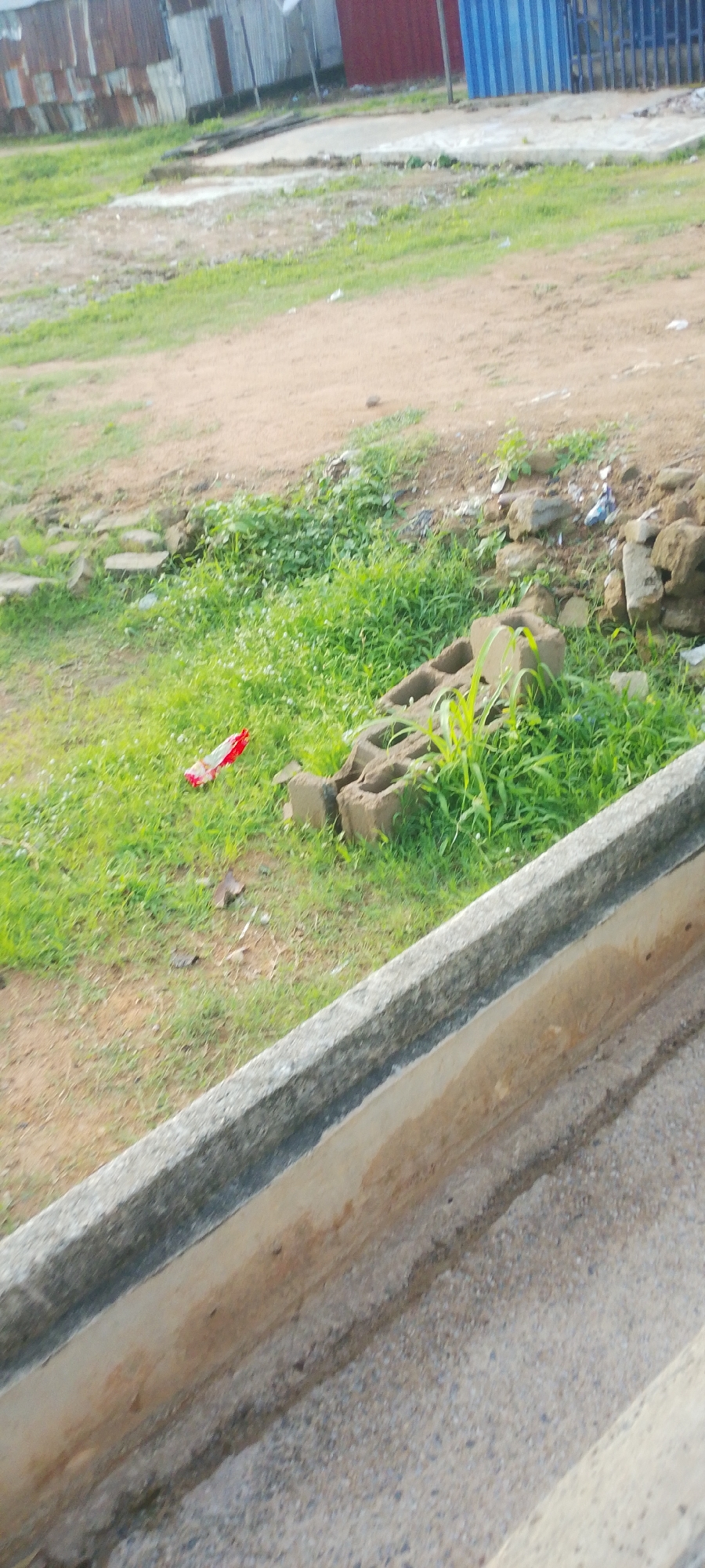 The image depicts an outdoor scene in Jalingo, Nigeria, likely during the daytime with overcast or bright conditions. The foreground features a concrete gutter with a rough texture and some discoloration, suggesting it is aged or has been exposed to the elements. Beyond the gutter, a grassy area is visible, with patches of dirt and scattered rocks. Some of the rocks appear to be piled up, possibly part of a rudimentary structure or debris.
In the grassy area, there are a few scattered objects. One prominent item is a red and white wrapped object lying on the grass. Further to the right, there are several concrete blocks, some stacked and others partially buried in the grass, with some green shoots growing around them, indicating they have been there for some time. The background shows a collection of buildings constructed with corrugated metal sheets and some sections of red and blue siding, which are typical of informal settlements or low-cost housing in many regions. The overall impression is one of an unkempt, possibly urban or peri-urban environment. There is no visible text in the image.