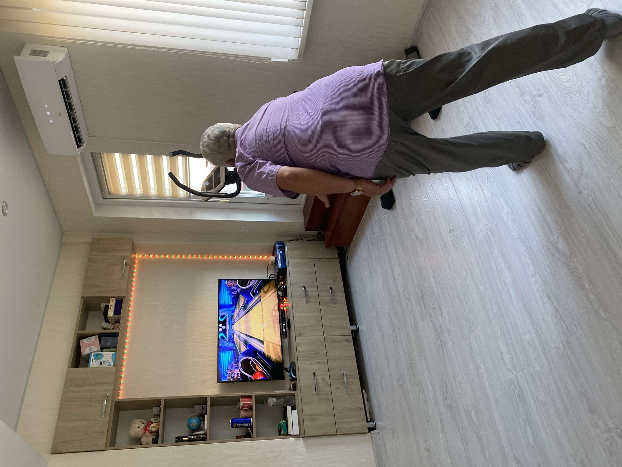 A person is standing in front of a television in a room and playing a bowling video game. The person is wearing a purple striped shirt and gray pants. The television is on a wooden cabinet with drawers. There is a shelf unit to the left of the television with various items on it. An air conditioner is mounted on the wall above a window with blinds. The floor is made of light-colored wood laminate. There is an elliptical machine in the background. The room appears to be a living room or recreation room.
