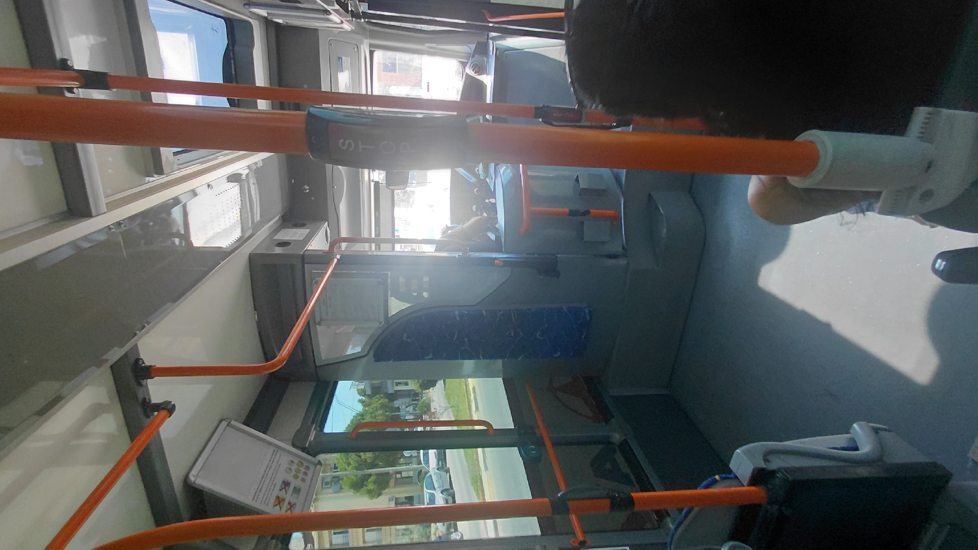The image shows the interior of a public bus, possibly in the Municipal Unit of Aegio, Greece. The bus features orange grab poles throughout the cabin, with grey and blue seating visible. Sunlight streams through the windows and a skylight, illuminating the otherwise dimly lit interior. In the foreground, a person's shoulder and arm are visible, suggesting they are a passenger. In the mid-ground, a stop request button is mounted on a pole, and the driver's area is partially visible with controls and a window looking out onto a street with cars and trees. The overall scene depicts the typical interior of a public transport vehicle.