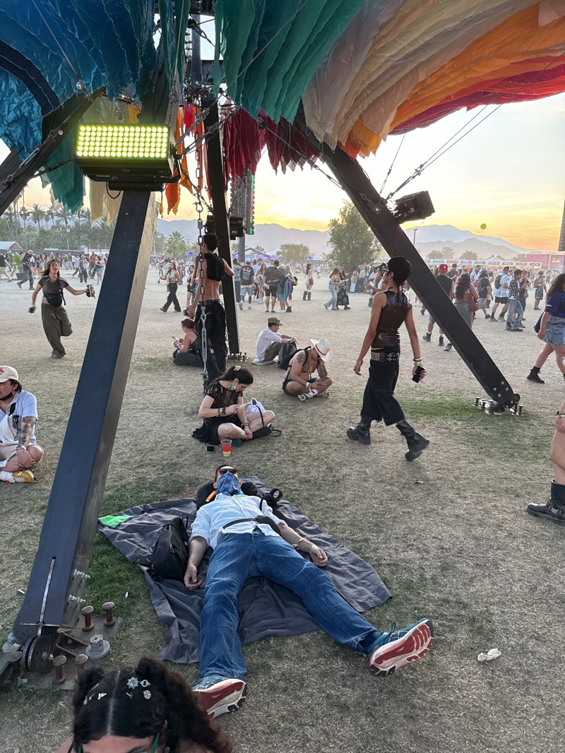 The image depicts a crowd of people at an outdoor event in Indio, United States. The location is a grassy area, possibly a park or festival ground, where some attendees are sitting or relaxing on the grass. In the foreground, a man lies on a blanket, wearing jeans, a light-colored shirt, and sneakers. He has a patterned covering over his face and appears to be sleeping or resting.
There are several other people scattered around the central figure. Some are sitting in small groups, and others are walking through the crowd. A large, multi-colored structure, possibly an art installation or shade canopy, dominates the upper part of the scene. There is bright lighting attached to the structure. The lighting is facing towards the ground. The sky is visible in the background, suggesting it is late afternoon or early evening. Palm trees can be seen in the distance, indicating a desert or arid environment.
