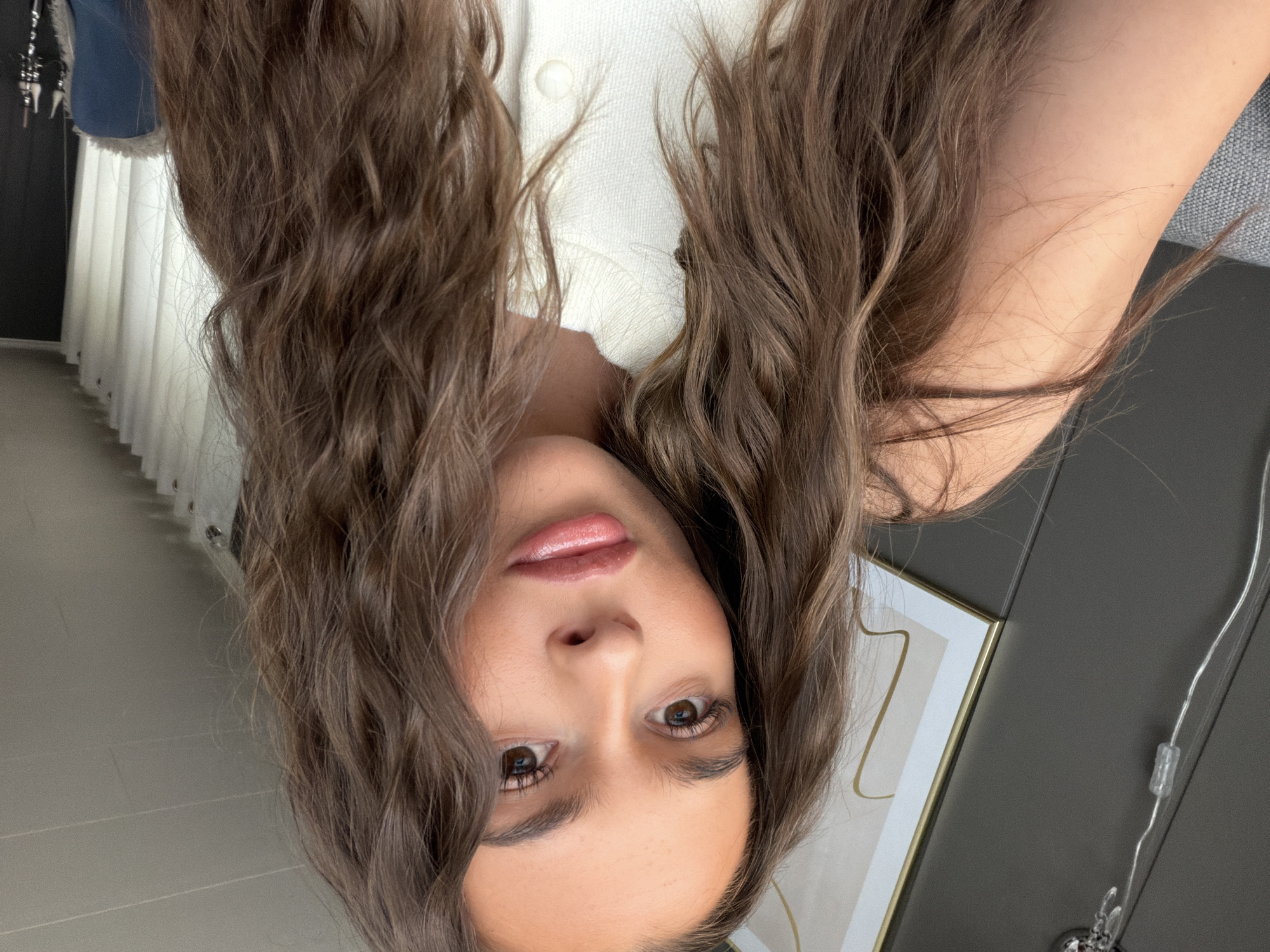 A young woman with long, wavy brown hair is taking a selfie indoors. She is wearing a white, button-up cardigan. Her makeup includes foundation, blush, eyeshadow, mascara, and lip gloss. In the background, a framed piece of abstract art with gold accents hangs on a dark gray wall. To the right of the frame, white curtains are visible. The image is a close-up shot, focusing on the woman's face and upper body. No specific location in Harstad, Norway is identifiable.