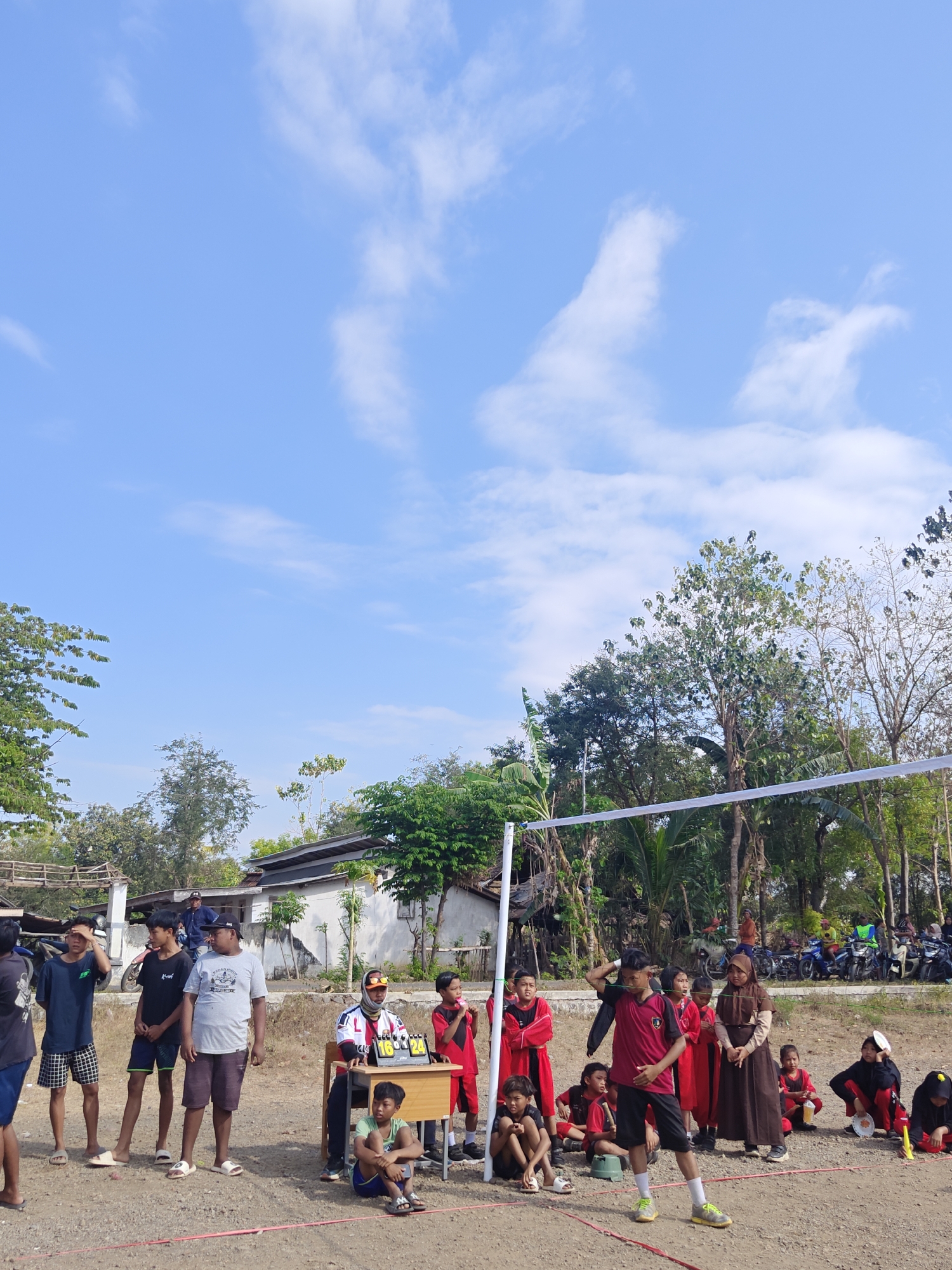 In Cermee, Indonesia, under a bright blue sky with scattered white clouds, a group of individuals is gathered on a sandy, outdoor court, likely for a sporting event. A white volleyball net stretches across the scene, supported by poles. To the left, several young males in casual attire, including t-shirts and shorts, stand and observe. A wooden table with a scoreboard displaying numbers 16 and 24 is positioned to the right, attended by a person in a dark hijab and an orange headscarf, wearing athletic attire. Beside the table, a young boy in a green shirt is seated on the ground. In the mid-ground, a group of children in red and black athletic uniforms are standing and sitting. A young person in a maroon and black athletic uniform is in the foreground, appearing to be in motion, possibly playing a sport. The background features houses, trees, and parked motorcycles.