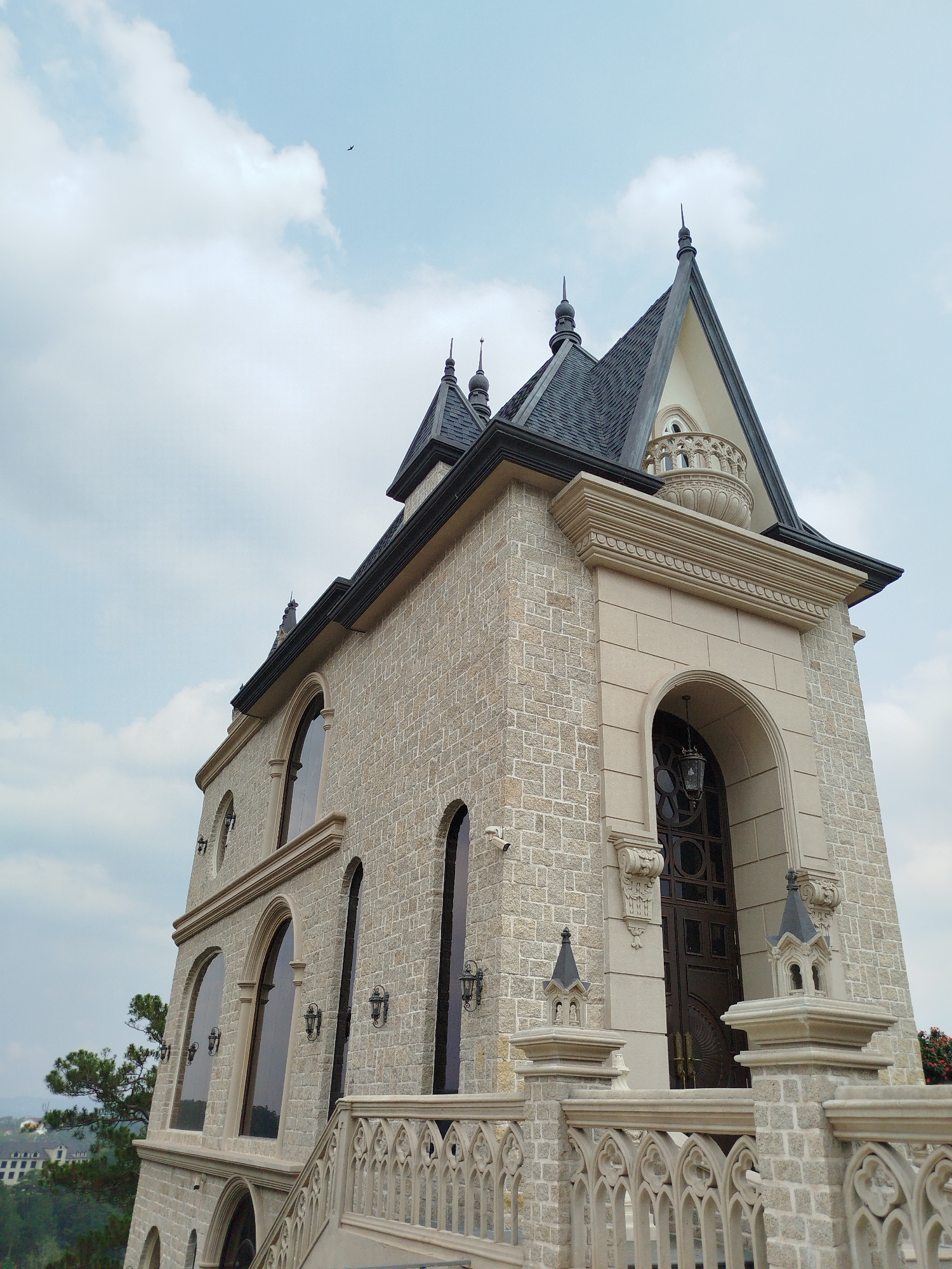 The image showcases a majestic multi-story building constructed in a neo-Gothic or castle-like architectural style, set against a bright, partly cloudy sky. The building features an exterior of light beige, rough-hewn stone blocks, giving it a textured and imposing appearance. Its roofline is characterized by steep, dark gray shingled roofs adorned with multiple pointed spires.
Numerous large arched windows, some with dark, intricate frames, pierce the stone facade. An ornate stone railing, featuring Gothic tracery patterns, is visible along a walkway or balcony, topped with decorative finials resembling miniature turrets. A grand arched entryway reveals a dark wooden door, with a decorative hanging lantern suspended within the arch. Wall-mounted sconces are placed at intervals along the building's exterior, and small security cameras are also visible. Near the peak of the main roof, a small, ornamental stone balcony adds to the building's intricate details.
In the background, green foliage, including a tall pine tree, and distant buildings nestled amidst hills suggest an elevated, scenic location. The sky is overcast with soft, diffused daylight, indicating daytime. A small, dark object, likely a bird or distant plane, flies in the upper left sky. The building's grandeur and European-inspired design are characteristic of locations like Da Lat, Vietnam. No people or visible text are present in the image.