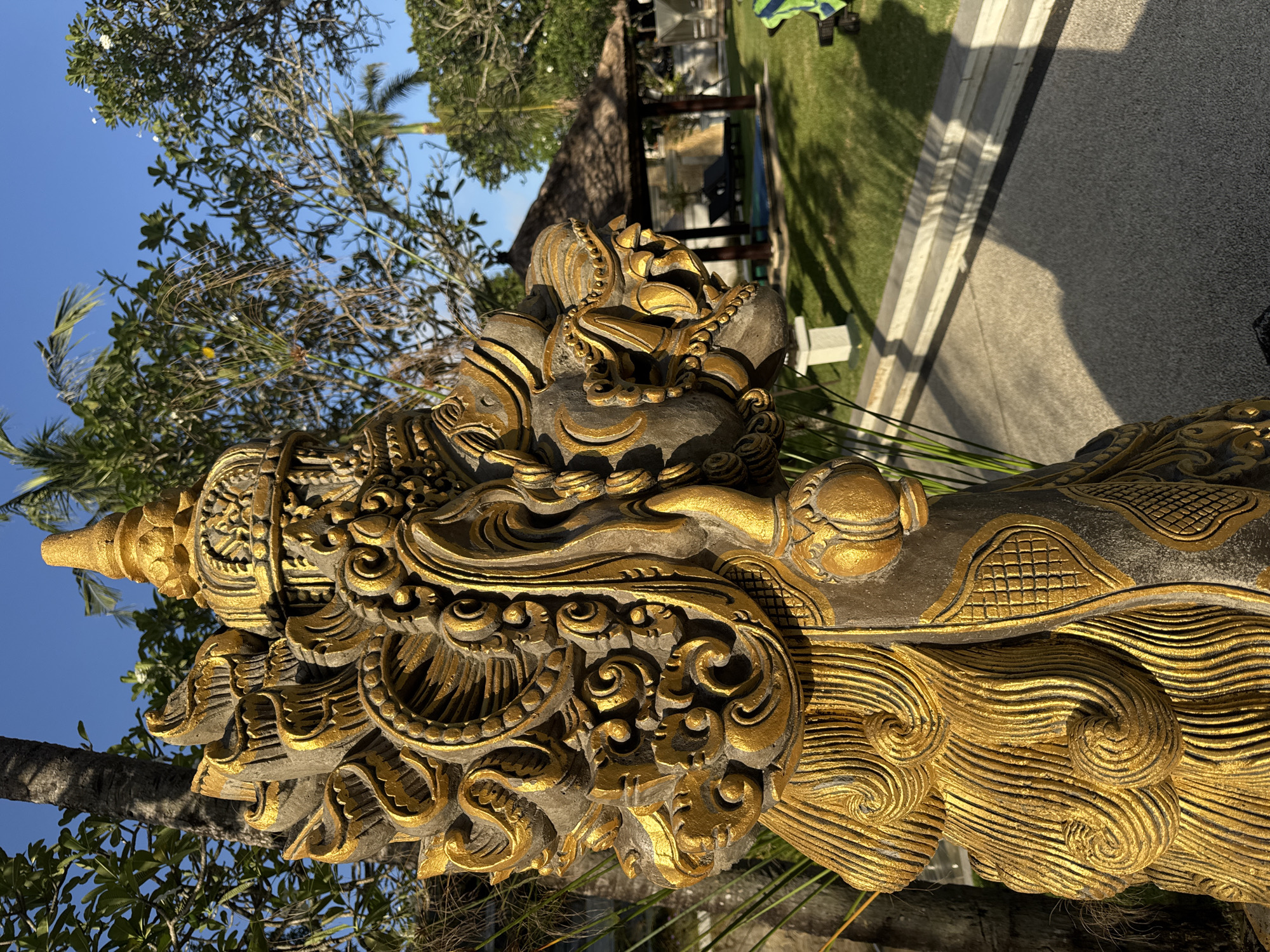 The image features a close-up of a heavily ornamented, gold-accented stone sculpture situated outdoors in Jimbaran, Indonesia. The sculpture's intricate details suggest a traditional Balinese style, with elaborate carvings and a weathered appearance. The sculpture is positioned in front of a paved area and a well-manicured lawn. A partially visible building with a thatched roof is visible in the background. The scene is captured under clear, bright daylight.