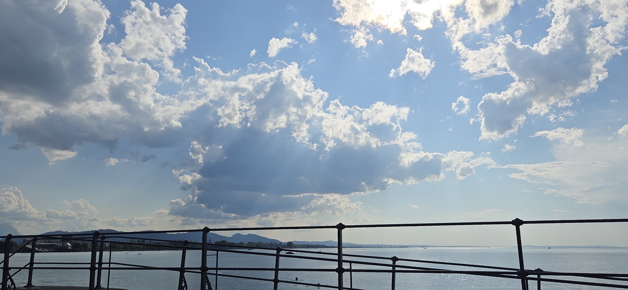 This is a wide shot taken from a railing overlooking a body of water. The sky is filled with dramatic clouds, with sunlight breaking through in places, creating visible sunbeams. The water is calm, reflecting the sky. In the distance, a low-lying landscape with hills and trees is visible, suggesting a natural setting. The railing in the foreground is dark and metallic, with a simple design. The scene appears to be a peaceful, scenic view, possibly from a promenade or a pier. The location is identified as Lochau, Austria.