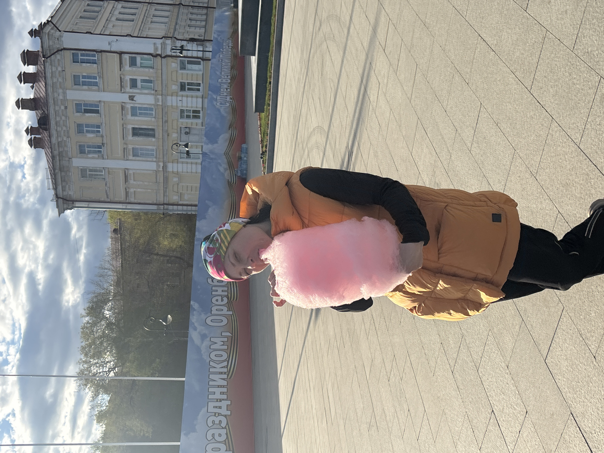 A person is eating pink cotton candy on a paved plaza in Orenburg, Russia. The person wears a yellow puffer vest over a black long-sleeved shirt, black pants, and black sneakers with white stripes. They also wear a colorful patterned head covering. Behind the person is a banner with text in Russian, including "Orenburg" and "Happy Victory Day". In the background is a building with multiple windows and a blue sky with clouds.