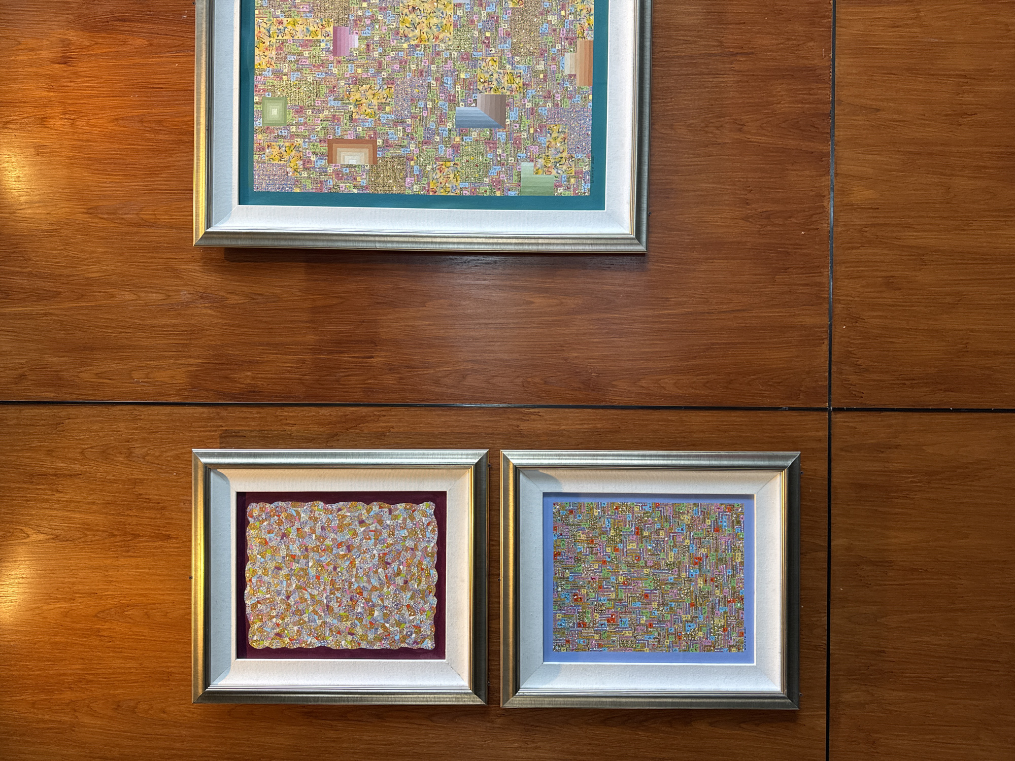 Three framed paintings are displayed on a wooden wall with vertical seams. The top painting is larger than the two below, which are placed side-by-side. The top painting has a gold frame with a white and teal inner border, and the painting itself features a mosaic-like pattern with squares and rectangles of various colors. The bottom left painting is framed in gold and white with a dark purple inner border, and displays an abstract pattern with dots and organic shapes in shades of orange, yellow, and white. The bottom right painting has a gold and white frame with a light blue border and a mosaic style painting with squares and rectangles in various colors. The wooden wall appears to be indoors and well-lit. The location is not discernible from the image, therefore no location can be derived.
