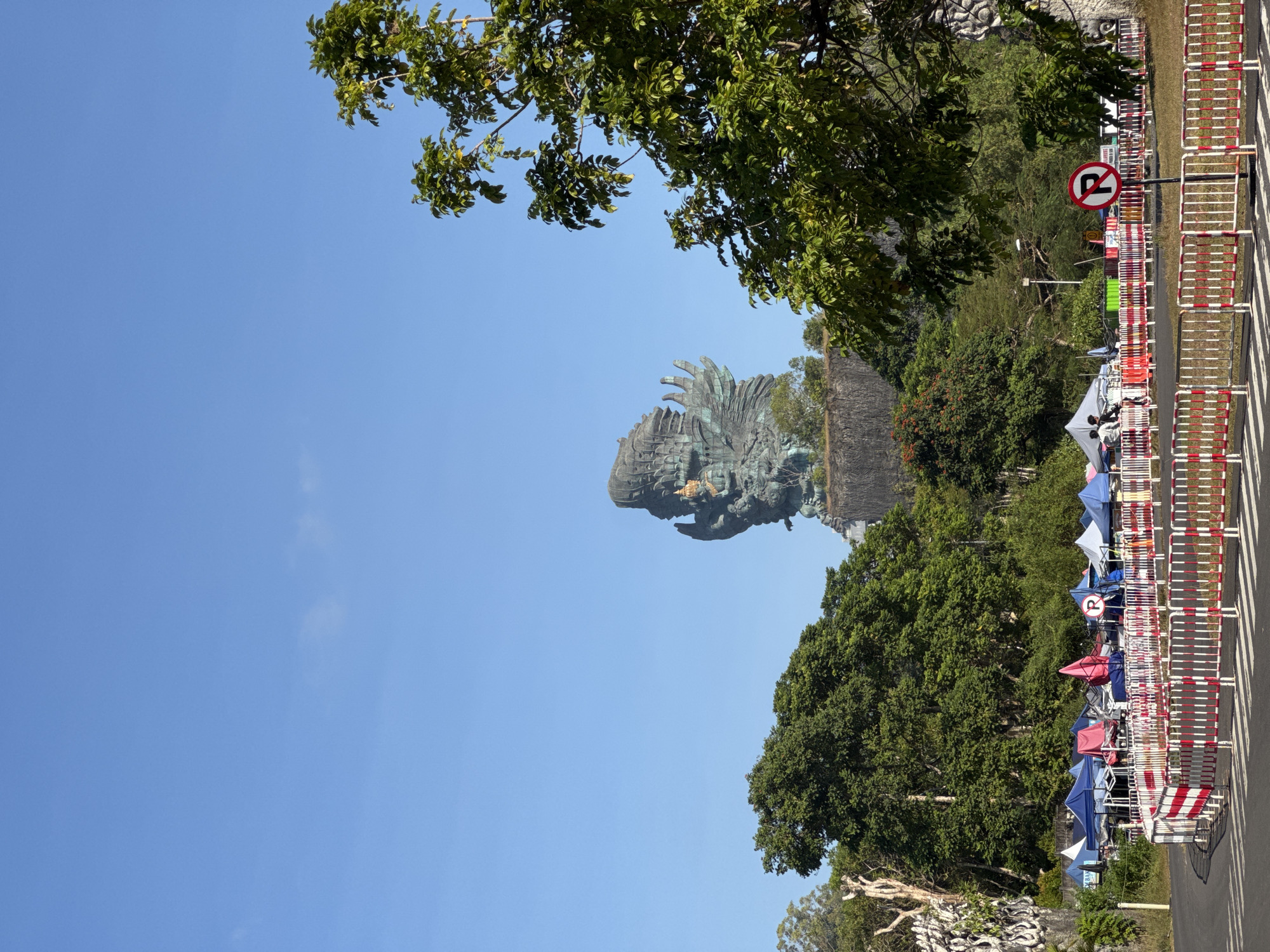 This image depicts a panoramic view under a clear blue sky in Kuta Selatan, Indonesia, featuring the monumental Garuda Wisnu Kencana statue. The colossal statue, appearing weathered bronze or green, dominates the upper-midground, rising above a dense band of varied green foliage and a rocky cliff face. The statue portrays the Hindu deity Vishnu atop the mythical bird Garuda, with a golden crown visible on Vishnu's head.
In the midground and foreground, a paved area or road curves gently to the left. Along the right side of this road, a continuous line of red and white striped metal barricades forms a barrier. Behind these barricades, numerous temporary market stalls or tents are set up, displaying various colors including blue, red, white, and green. Several indistinct figures can be seen near these tents, suggesting activity. A "No Parking" sign (a red circle with a diagonal line over a black 'R') is prominently displayed on a pole just behind the barricades on the right side. Lush green trees with dense foliage frame both sides of the foreground and midground, with one large tree prominent on the right. The overall scene is brightly lit, indicating clear daytime conditions.