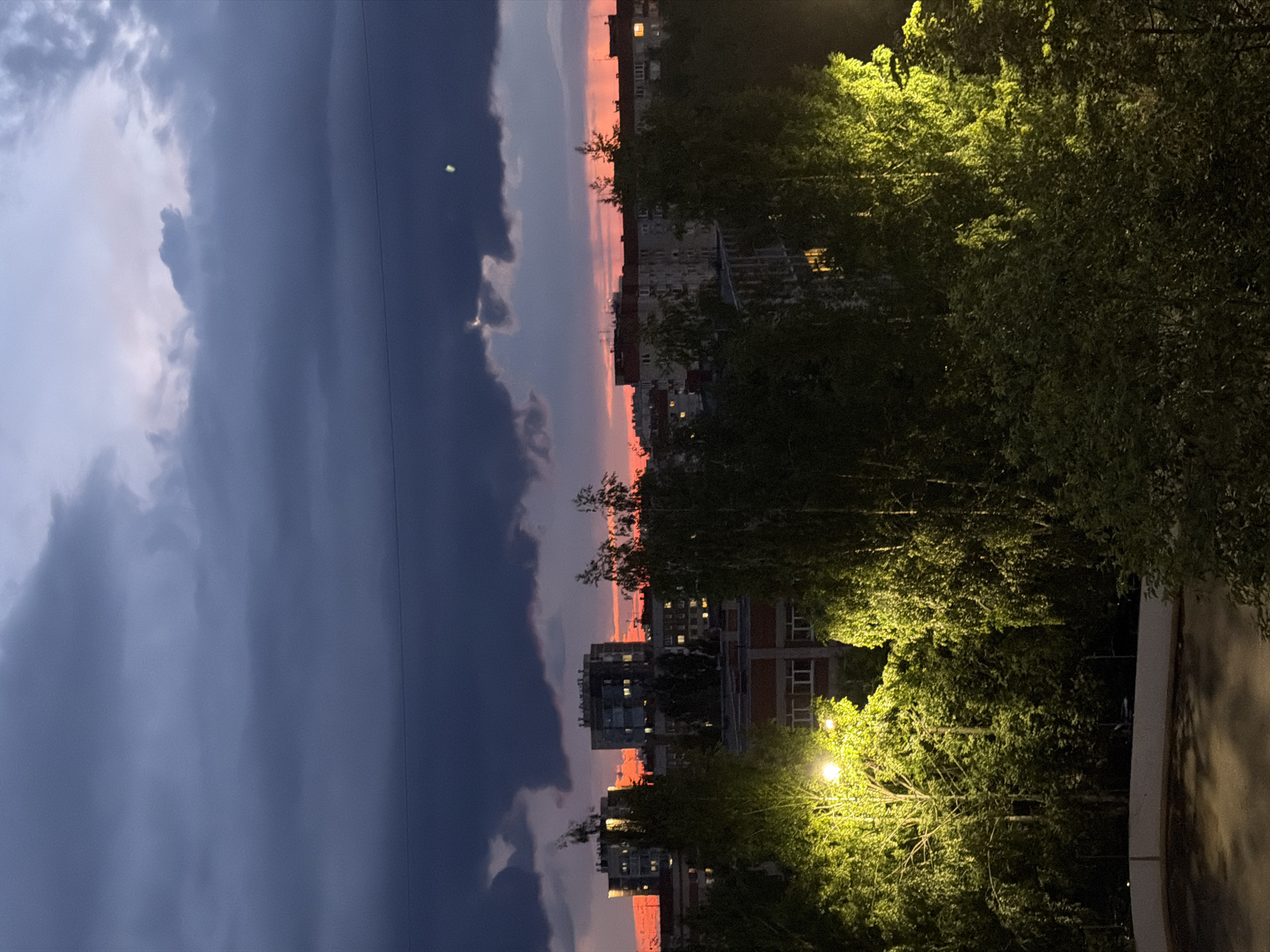 The image captures a twilight cityscape in Izhevsk, Russia, characterized by dramatic cloud formations and a vibrant sunset. The sky is dominated by dark, heavy clouds, with a strip of fiery orange and pink light breaking through near the horizon, indicating the setting sun. Silhouetted against this backdrop are multi-story residential buildings, some with illuminated windows suggesting occupancy. In the foreground, lush green trees are illuminated by streetlights, casting a warm, artificial glow on their foliage. The scene appears to be from a residential area, possibly a balcony or elevated viewpoint overlooking a park or green space bordered by apartment blocks. No people are visible in the image. The overall impression is one of an urban environment transitioning from day to night under a moody sky.