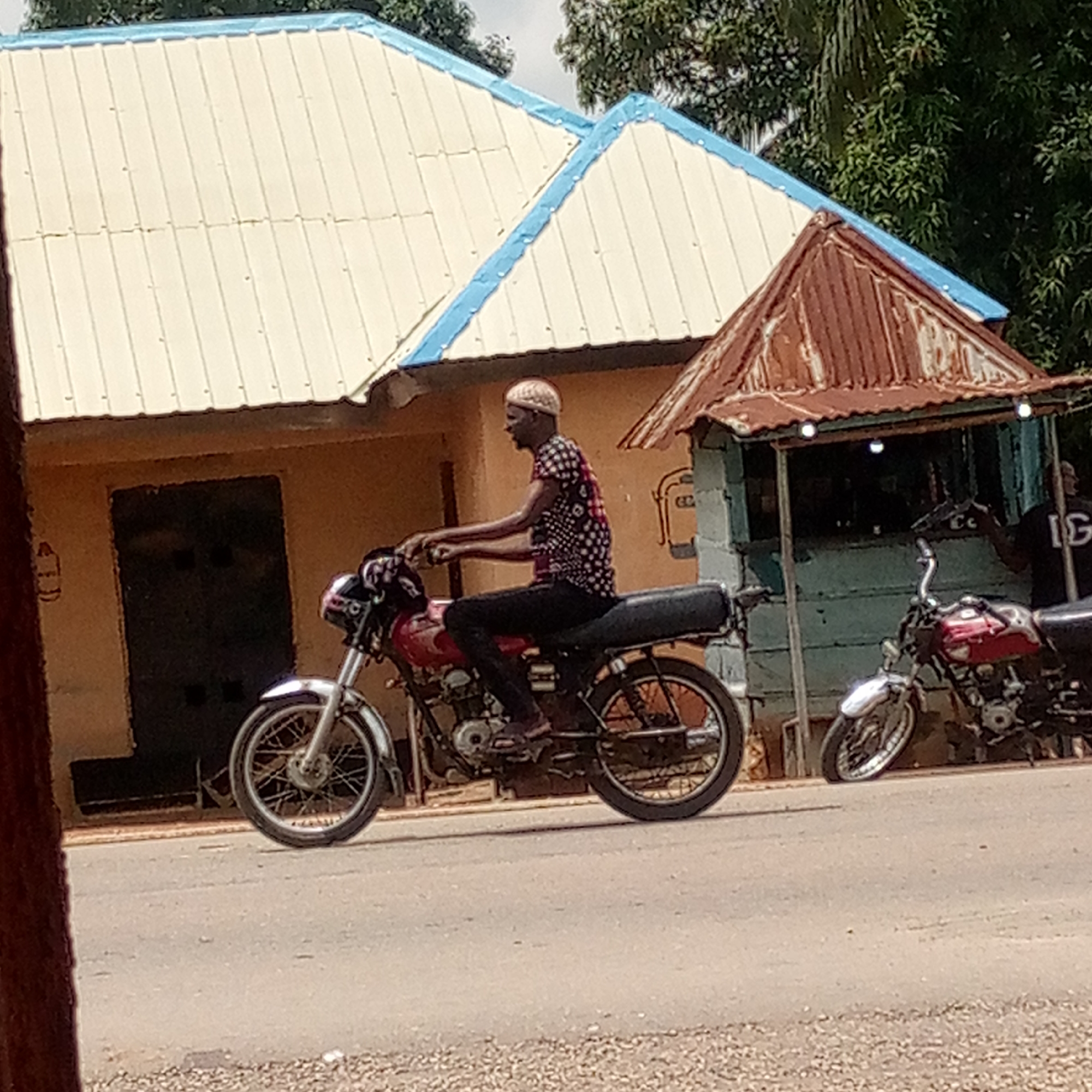 This outdoor daytime image, set in Mararaba, Nigeria, features a man riding a red motorcycle on a paved road. He is depicted in profile, facing left, wearing a light-colored head covering or short hair, a dark patterned short-sleeved shirt with white dots, dark trousers, and sandals. His hands are on the handlebars and his feet are on the footrests, suggesting active riding.
To the right of the rider, another red motorcycle is parked, and a third motorcycle is partially visible on the far right. In the background, a light-colored building with a distinctive cream-colored, corrugated metal roof accented with light blue trim is visible. This building has a dark-colored door. Next to it, a small, light-blue painted wooden stall or kiosk stands, featuring a rusty, corrugated metal roof and small lights fixed to its canopy. A person wearing a black t-shirt with white letters spelling "IG" is partially visible behind this stall. Lush green foliage from trees is seen in the upper background. The road surface is composed of asphalt with areas of loose gravel. The scene depicts a common street or roadside environment, typical of a bustling local area.