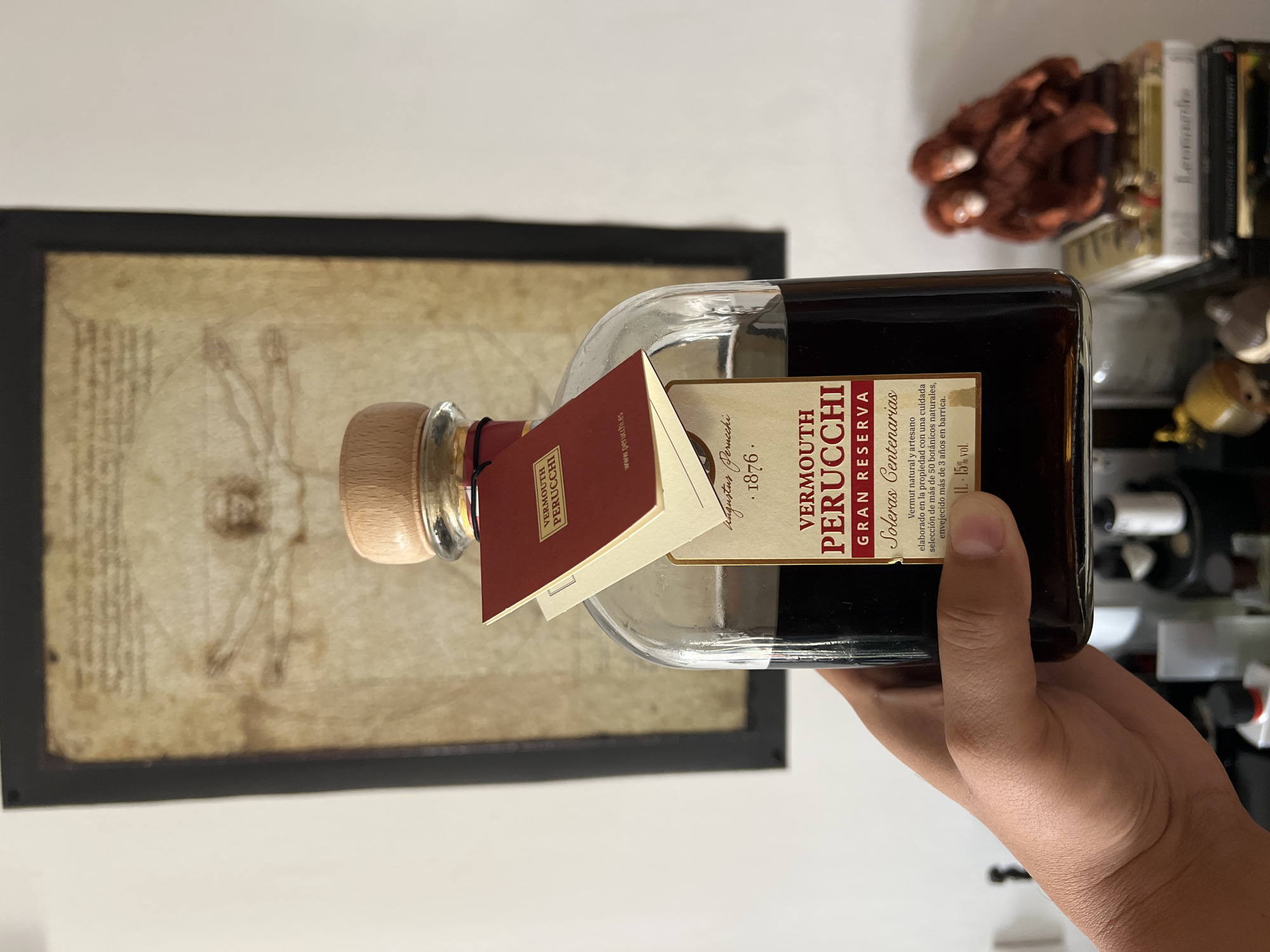 A person's hand holds a clear glass bottle of dark red vermouth labeled "VERMOUTH PERUCCHI GRAN RESERVA". The bottle has a wooden cork stopper and a red tag attached to its neck. Behind the bottle, a framed drawing of Vitruvian Man hangs on a white wall. To the right of the bottle, on a shelf, are several books and a small decorative sculpture of two figures. The scene is indoors, likely in a home, as suggested by the personal items and decor. The image is taken from a slightly low angle, looking up towards the bottle and the artwork. The lighting appears to be natural, coming from the right side of the frame, casting soft shadows. There is no information in the image to suggest a location in Mollet del Vallès, Spain.