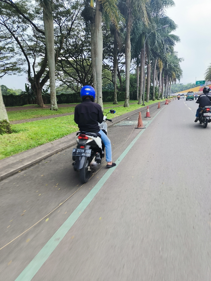 The image shows a man riding a black motorcycle on a paved road in Padalarang, Indonesia. The man is wearing a blue helmet and is in the process of overtaking another motorcycle. Both motorcycles are on the right side of the road, which is marked with a green line. There is a line of palm trees on the left side of the road, and there is green grass and vegetation behind the palm trees. There are orange traffic cones on the right side of the road, and there is a road sign in the distance on the right side of the road. The image was taken during the daytime. The weather appears to be clear. The image was taken in Padalarang, Indonesia. The specific location is near the intersection of Jalan Raya Padalarang and Jalan Cileunyi, as indicated by the coordinates.