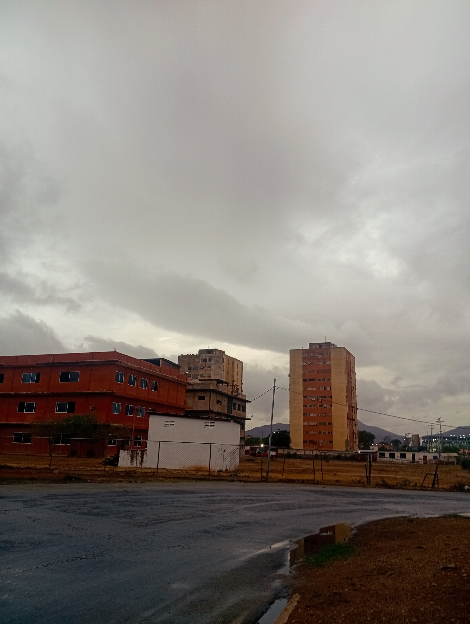 The image depicts a cityscape with several buildings under a cloudy sky in Juan Griego, Venezuela. In the foreground, a wet asphalt road curves towards the right, with patches of dry brown earth and sparse vegetation along its edge. A chain-link fence runs horizontally across the mid-ground.
To the left of the fence is a low-rise, orange brick building with numerous rectangular windows. Adjacent to it and slightly behind is a white building with some visible ventilation or utility openings. Further into the background, two taller buildings dominate the scene. One is a multi-story building with an orange-brown facade and many windows, appearing to be a residential or office tower. To its left, partially obscured, is another older, more weathered high-rise building. In the distance, the outlines of mountains are visible against the overcast sky. The overall atmosphere is subdued due to the cloudy weather. No people or animals are visible in the frame.