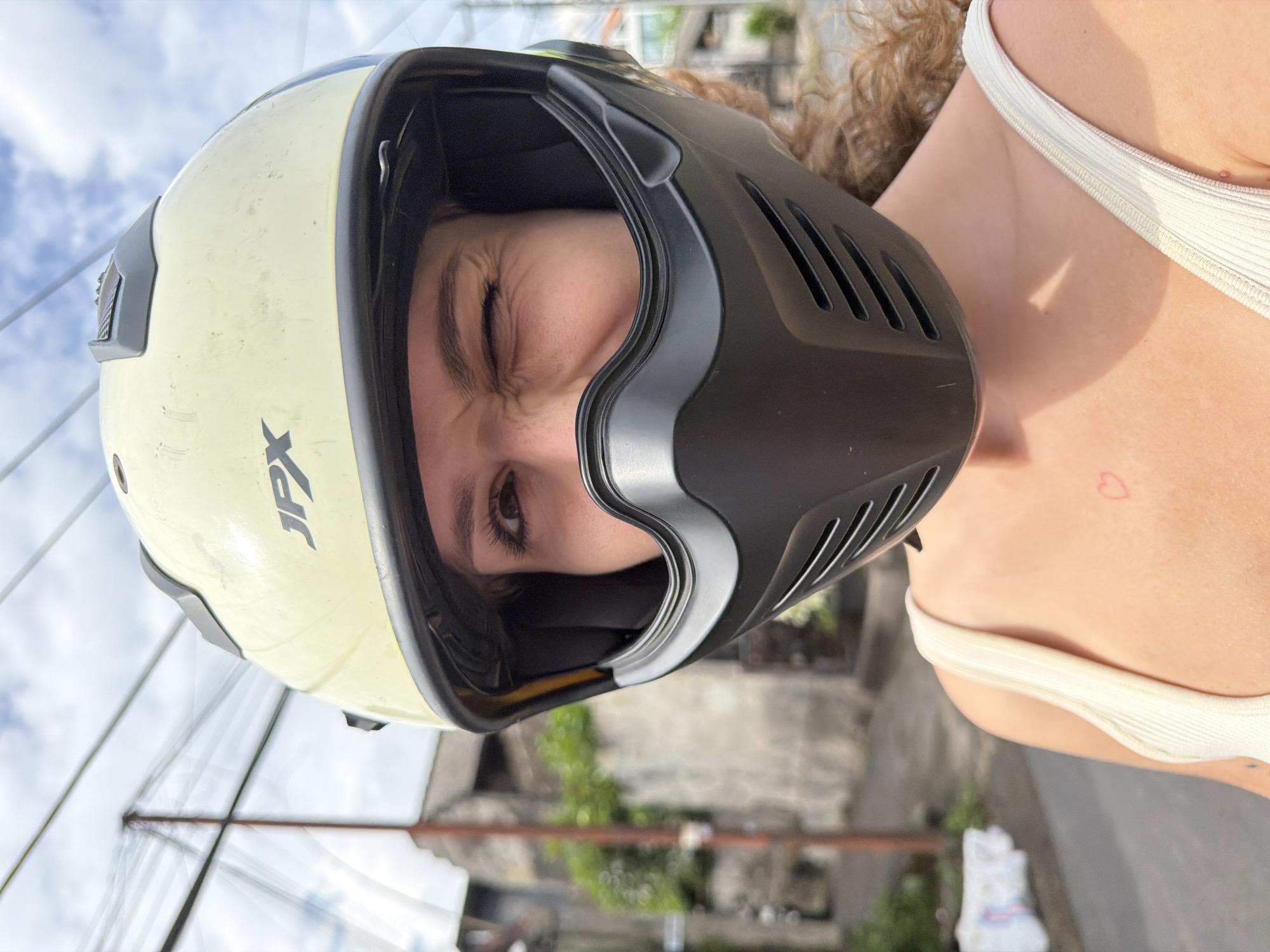 A young woman wearing a pale yellow motorcycle helmet with a black face mask is in the city of Mengwi, Indonesia. She is winking at the camera. The helmet has the "JPX" logo on it. The woman is wearing a cream-colored tank top. A faint pink heart tattoo is visible on her upper chest. The background shows a street with buildings, trees, and overhead power lines. The sky is partly cloudy.