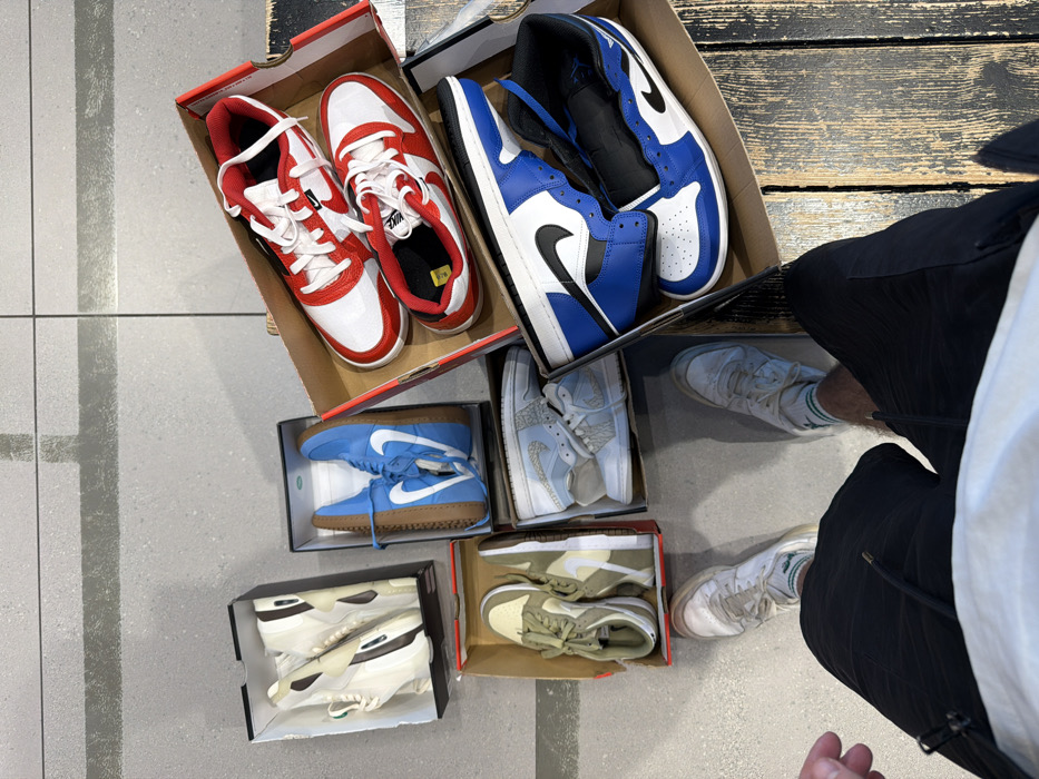 An overhead photograph depicts the lower body of an individual standing in a retail environment in Rishon LeZion, Israel, surrounded by multiple open shoe boxes containing athletic footwear. The person is wearing black shorts, white socks with green stripes, and a pair of white sneakers. On the floor, six open shoe boxes are arranged around the person's feet. These boxes contain various Nike brand sneakers: a pair of cream/off-white chunky trainers, a pair of olive green low-top sneakers, light blue Air Force 1s with a gum sole, red and white low-top sneakers (one with a yellow size sticker), light grey/white textured sneakers, and blue, white, and black Air Jordan 1 high-top sneakers. The flooring transitions from light grey square tiles on the left to dark, distressed wooden planks on the right. The scene suggests a shopping activity in a shoe store.