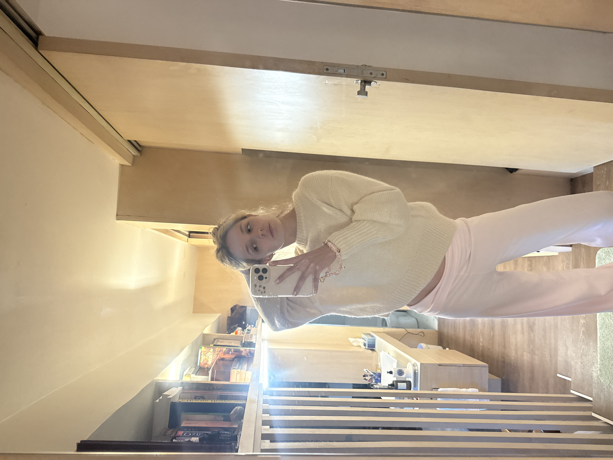 A woman is taking a mirror selfie in a room that appears to be a modern apartment or house. She is wearing a cream-colored, oversized knit sweater and pale pink wide-leg pants. Her blonde hair is pulled back in a messy bun, and she is holding a white smartphone in her right hand, which is adorned with a pink beaded bracelet. Her left hand is resting on the back of her head.
The background of the reflection shows a bookshelf filled with books on the left, a bathroom vanity with sinks and bottles on a counter, and a wooden sliding door on the right. The flooring is wood-look laminate. The lighting is warm and ambient, suggesting indoor lighting. The scene is devoid of any specific landmarks or clues that would place it in Ciudad de México, Mexico; it is a generic interior space.