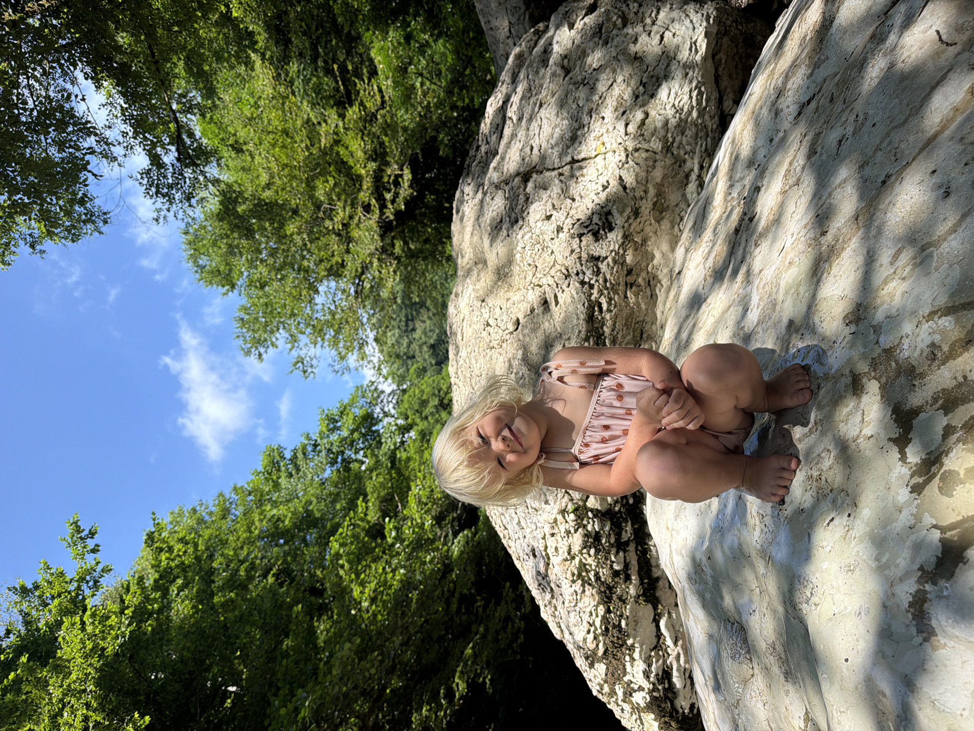 A young girl with blonde hair is sitting on a large, textured rock, which appears to be wet. She is wearing a pink swimsuit with a polka dot pattern. The girl is smiling and looking towards the camera. Her legs are drawn up towards her chest, and her hands are clasped together.
The scene is outdoors, with lush green trees and a bright blue sky with white clouds visible in the background. Sunlight is casting shadows on the rock, suggesting it is daytime. The overall impression is one of a pleasant, sunny day in a natural setting. The girl's expression and posture convey a sense of happiness and comfort. There are no visible text elements in the image. The location context provided is Khostinskiy, Russia.
