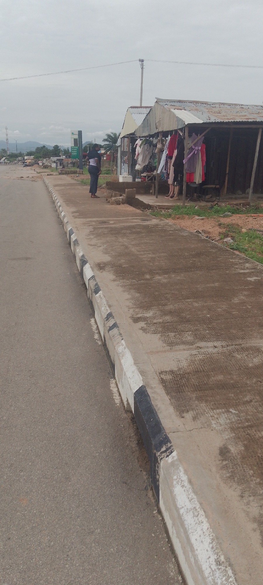 The image depicts a roadside scene in Jalingo, Nigeria, with a focus on a small market stall and the adjacent paved walkway and road. In the foreground, a gray asphalt road runs diagonally from the bottom left to the middle of the frame. A concrete curb with alternating black and white painted sections borders the road and separates it from a concrete walkway.
On the walkway, a person is standing, facing away from the camera and towards a market stall. The person is wearing a dark-colored top and pants. The market stall, constructed with corrugated metal roofing and siding, is positioned further back on the walkway. It displays various articles of clothing hanging on a rack. Beyond the stall, the environment appears somewhat rural or semi-urban, with some vegetation, distant hills, and what appears to be a gas station sign visible in the background. The sky is overcast.