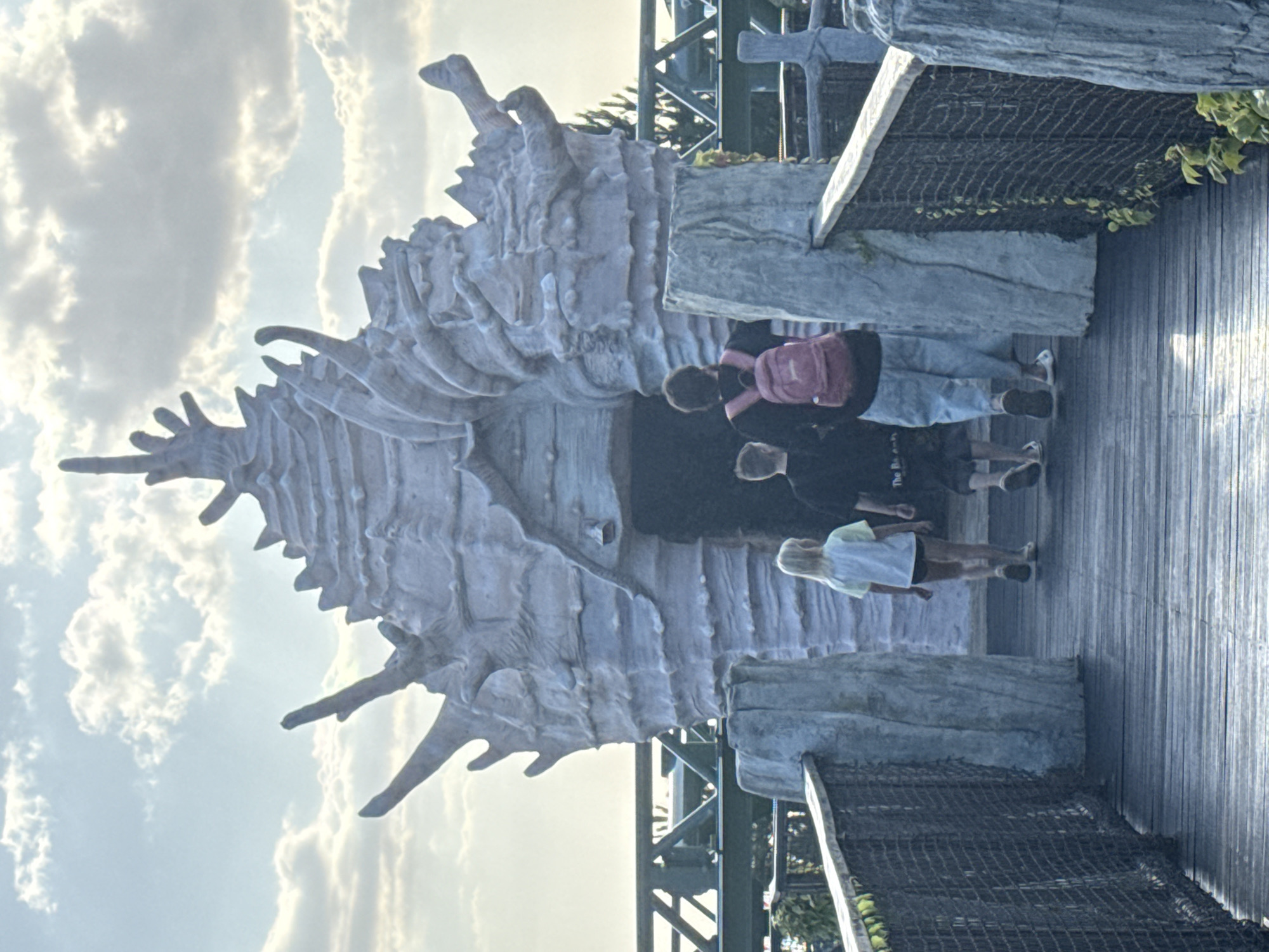 Three people are walking on a wooden walkway towards an entrance shaped like a large, pale shell. The person in front is wearing a dark shirt and light blue pants and has a pink backpack. To their left, a child with blonde hair wears a light-colored shirt and dark shorts. To the right of the person in the blue pants, another child in a dark shirt and dark shorts walks. The walkway is lined with railing made of wire mesh and light gray, textured pillars. In the background, a green metal structure, possibly a roller coaster track, is visible against a cloudy sky. The shell-shaped entrance is ornate, with spikes and ridged textures. The overall setting appears to be an outdoor attraction.