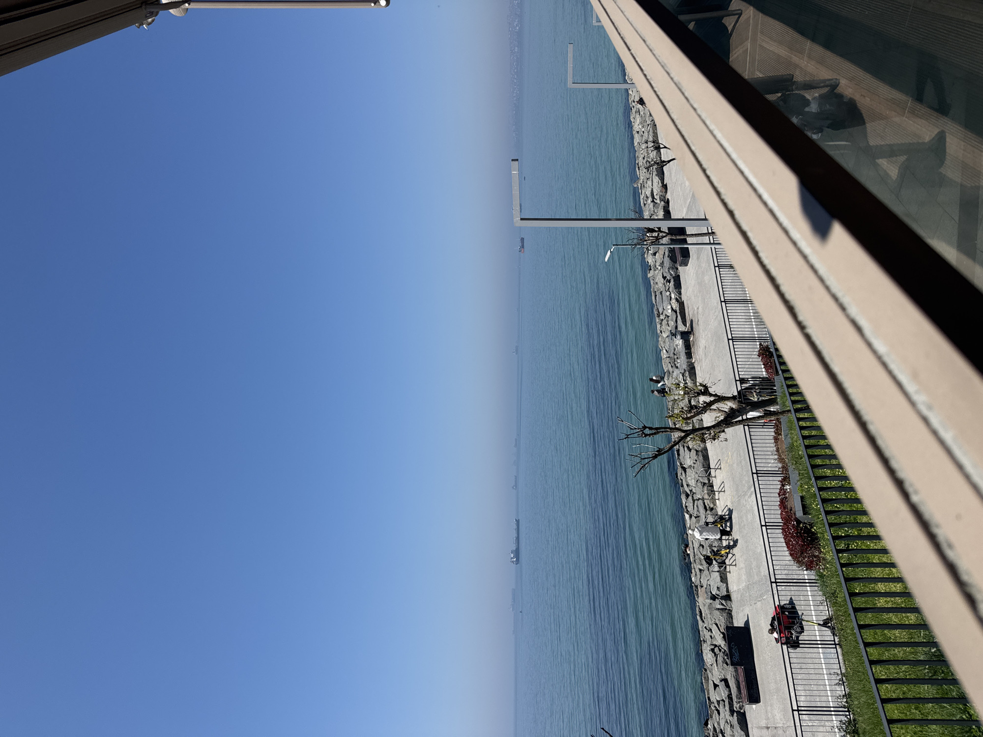 An oblique, long-shot shows a coastal view in Bakırköy, Turkey. Azure sky fills the left frame, meeting a blue-green body of water at a distant horizon punctuated by indistinct ships. A concrete promenade follows the rocky shoreline, lined with metal railings. A variety of benches, lampposts and small trees are interspersed along the promenade. A man, wearing a white top, can be seen sitting down next to a stone path that separates the grass from the pathway. A red vehicle is parked along the walkway. The right frame is filled by a building facade. Reflections of people can be seen in the glass windows.
