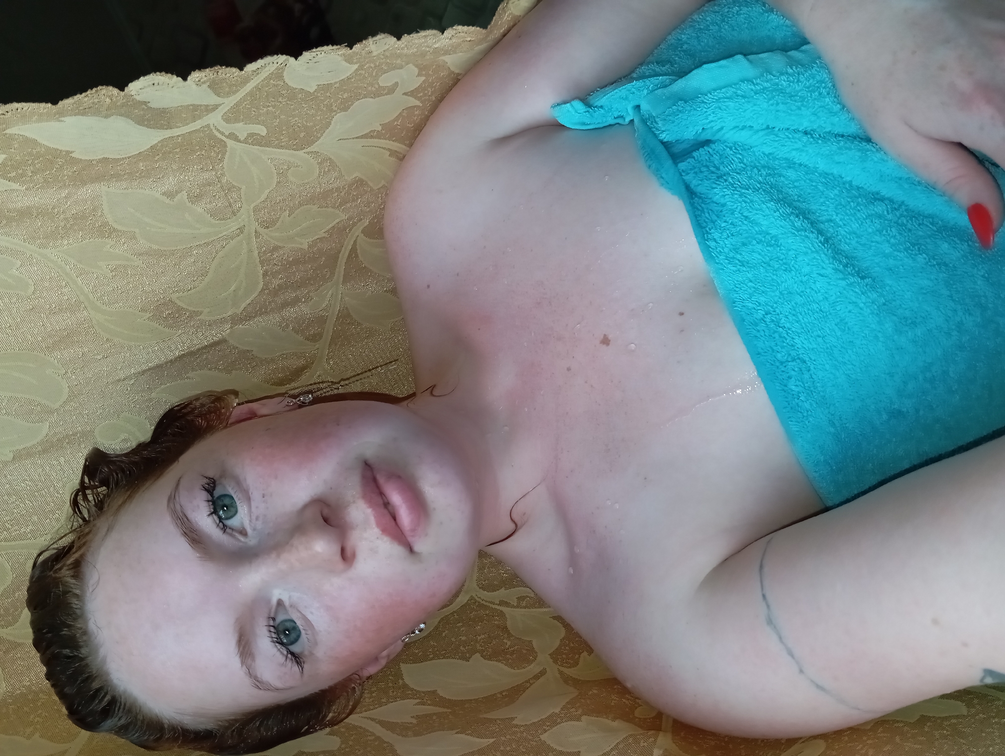 A fair-skinned person with wet brown hair and blue eyes is lying on a gold patterned surface. They are partially covered with a turquoise towel, held in place by a hand with red nail polish. The person appears to be slightly damp. A tattoo is visible on the person’s left arm. The location is undetermined. The reference to Yurgamysh, Russia is not supported by the image.