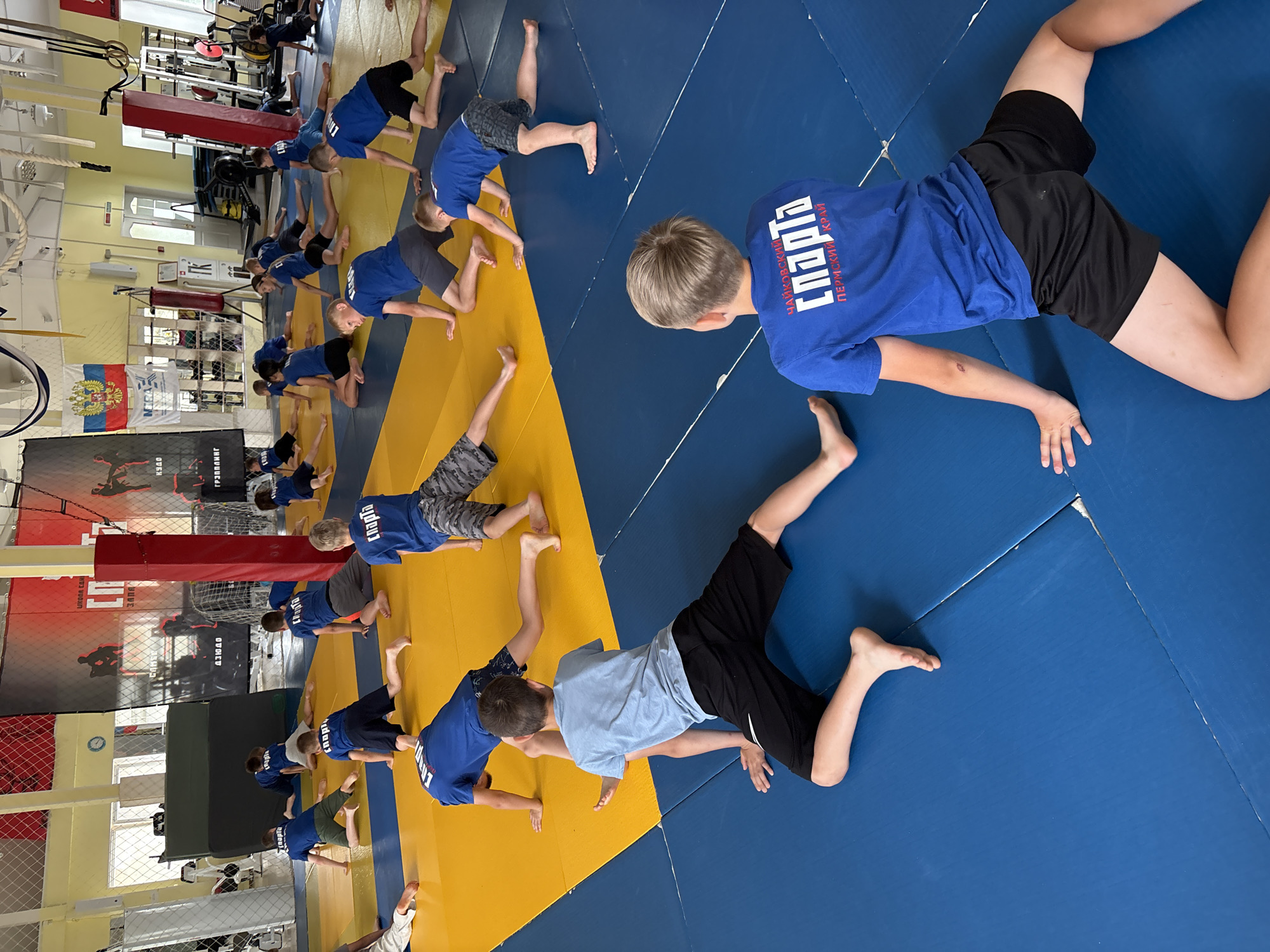 The image depicts a group of young boys in athletic attire engaged in a warm-up or stretching exercise on a blue and yellow mat in what appears to be a sports hall in Chaikovsky, Russia. The boys are wearing blue t-shirts with "СПАРТА" (Sparta) logo and black shorts. The environment includes wrestling mats, a punching bag, gym equipment, and flags, suggesting a martial arts or wrestling training facility. The boys are positioned in various yoga-like poses, indicating a structured physical activity session. The overall scene is one of a sports training environment with active participants.
