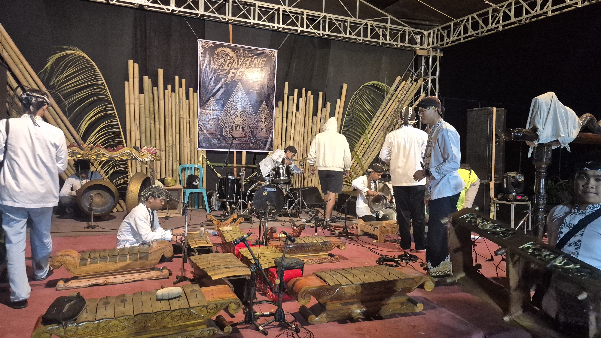 This image captures an outdoor performance scene at the "GAYBING FESTIVAL" in Bumen, Indonesia. The stage is decorated with bamboo stalks and palm fronds, creating a naturalistic backdrop. In the foreground, a collection of gamelan instruments, including metallophones and gongs, are arranged on a red carpeted floor. Several individuals are present, some appearing to be musicians. One man in a white shirt and black pants is seated near a gamelan, holding a mallet. Another man in a white hoodie stands near a drum set, while others in traditional attire are visible throughout the scene, suggesting a cultural music performance. The lighting suggests it is evening or nighttime.