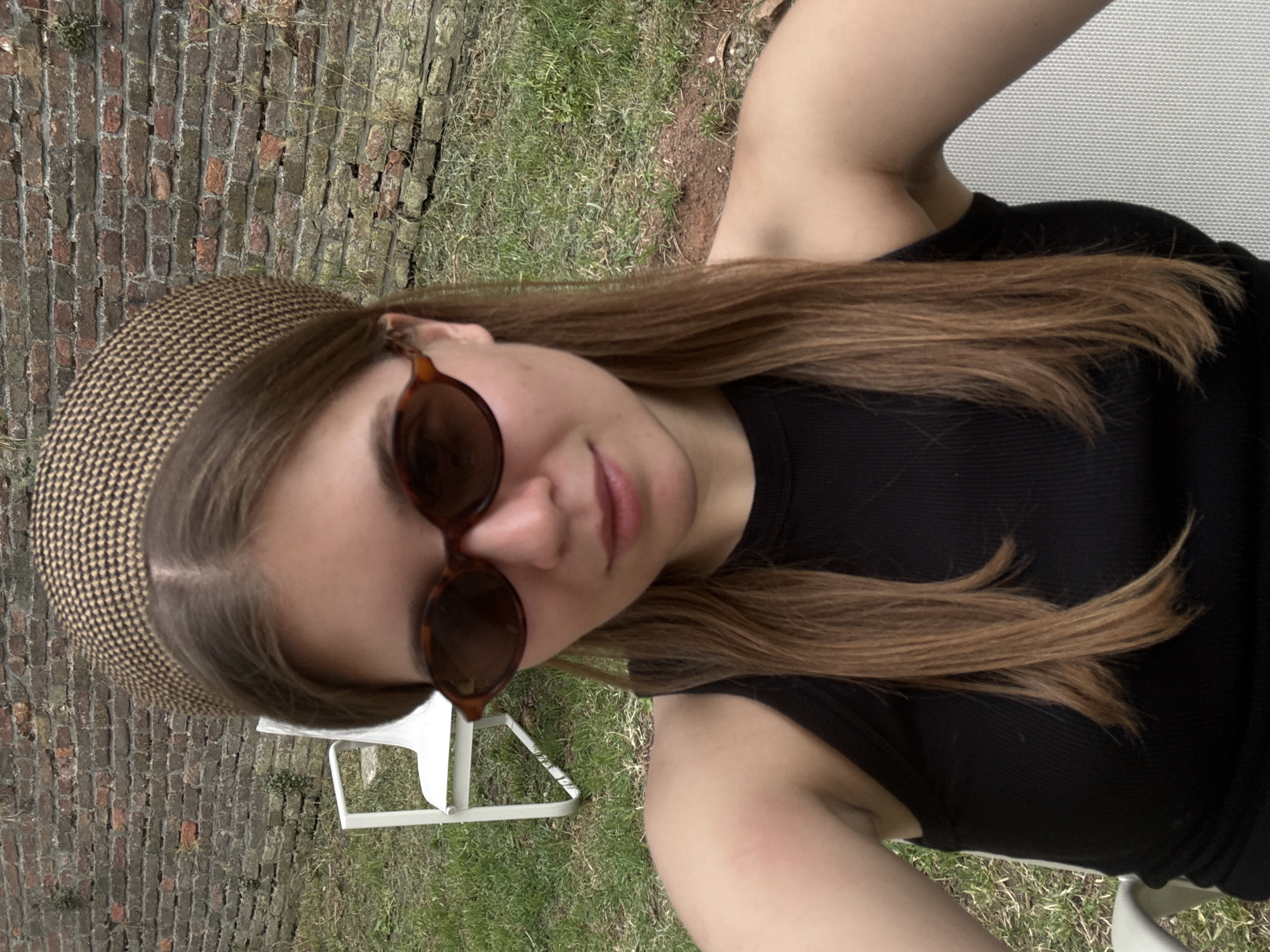 A medium-shot photograph shows a young woman with light brown hair wearing sunglasses and a patterned headband, seated outdoors. She is wearing a black tank top. The background consists of a brick wall, patches of grass, and a glimpse of a white chair. The location cannot be determined to be Belgrade, Serbia.