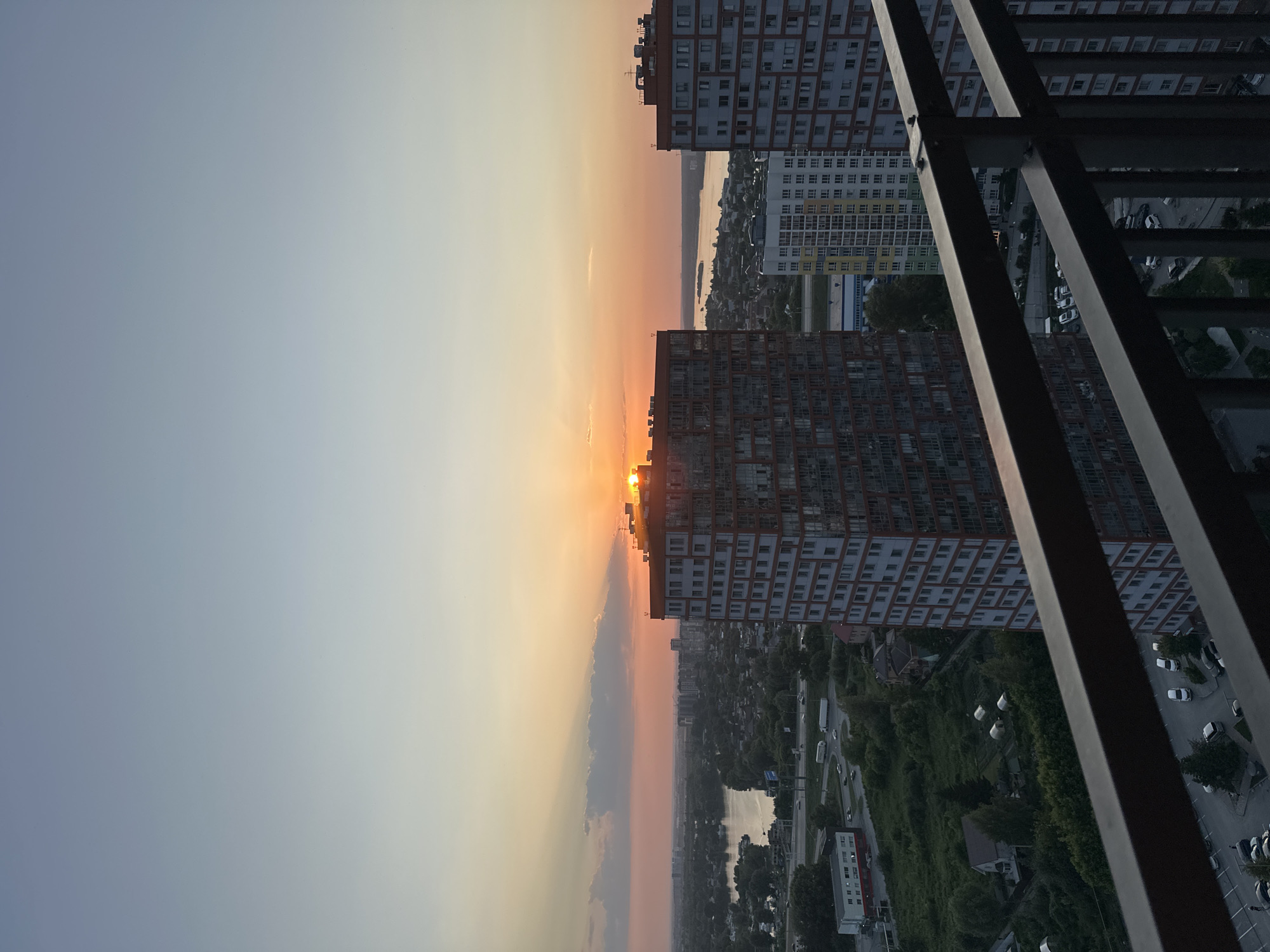 This is a photograph taken from a high vantage point, likely a balcony or window, looking out over a cityscape at sunset. The image is oriented vertically, with the sky and horizon occupying the left half and the city, buildings, and foreground railing occupying the right.
The sky is filled with the warm colors of sunset, transitioning from a soft orange and yellow near the horizon to a pale blue and grey overhead. Clouds are present, catching the light of the setting sun.
Dominating the midground are several tall, multi-story buildings. One building, on the left, has a facade of red and white blocks, with many small windows. To its right, another building is predominantly white and grey, also with numerous windows, and some sections with yellow and green accents. Further into the distance, more buildings are visible, creating a dense urban environment.
Below the buildings, a landscape of trees, roads, and smaller structures is visible. Cars are parked along streets and in parking lots. A body of water, possibly a river or lake, can be seen in the distance, reflecting the sky.
In the immediate foreground, a dark metal railing runs diagonally across the right side of the frame, obscuring some of the view and adding a sense of depth.
The time of day is clearly sunset, indicated by the colors in the sky and the low angle of the sun, which is partially obscured by a cloud and a building. The weather appears to be clear with some cloud cover. No people are discernibly present in the image. The overall mood is peaceful and serene, with the warm hues of the sunset creating a tranquil atmosphere. No text is visible in the image.