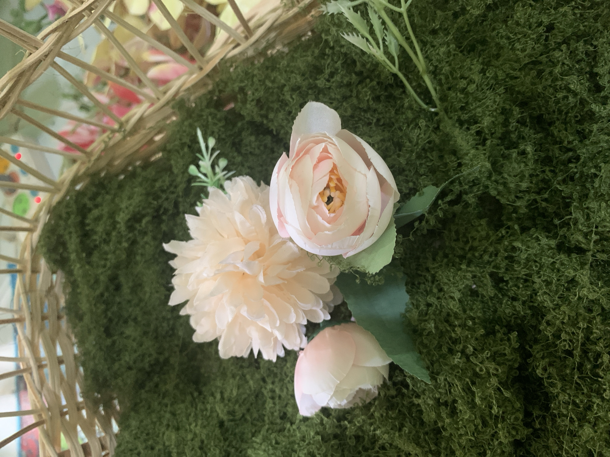 A close-up, overhead shot shows a collection of artificial flowers arranged on a bed of green moss. In the background, a woven basket made of light brown reeds is partially visible, containing blurred colorful items. The flowers consist of a large, pale peach chrysanthemum-like bloom, a light pink rose that is partially open with yellow and orange hues at its center, and two tightly closed buds in a similar light pink color. Green artificial leaves are attached to some of the flowers, and a few sprigs of greenery are scattered among them. The scene appears to be set up for decoration or display. No people are present in the image. The location is not specified in the provided context.