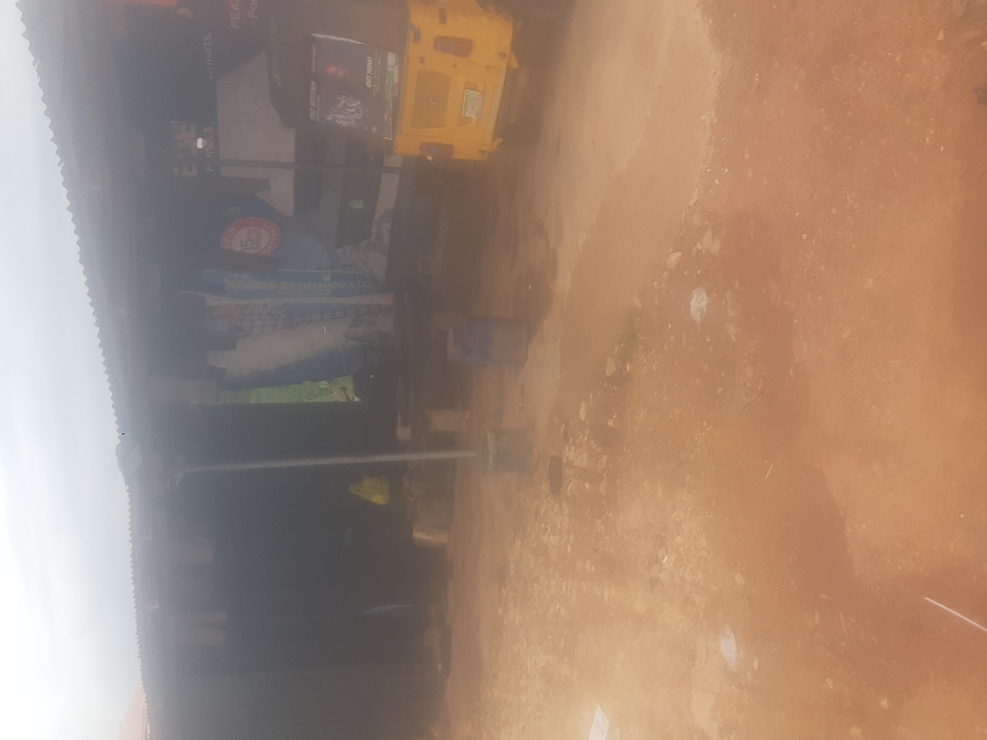 The image captures a dimly lit outdoor scene, likely on a dusty road in Enugu, Nigeria. The foreground is dominated by reddish-brown dirt and scattered stones. In the background, a structure with a corrugated metal roof appears to be a shop or market stall. A yellow auto-rickshaw, a common mode of transport in Nigeria, is parked to the right. The rickshaw has a banner on its rear that reads "THE RETURN" and "OUT NOW!". Behind the rickshaw, a large poster with text is visible on the wall of the structure. To the left, a dark interior of the structure is seen, with a yellow plastic bag hanging and some unidentifiable objects. The overall impression is one of a utilitarian, possibly commercial, area in a developing urban environment, with limited visibility due to the lighting conditions and potential dust in the air.