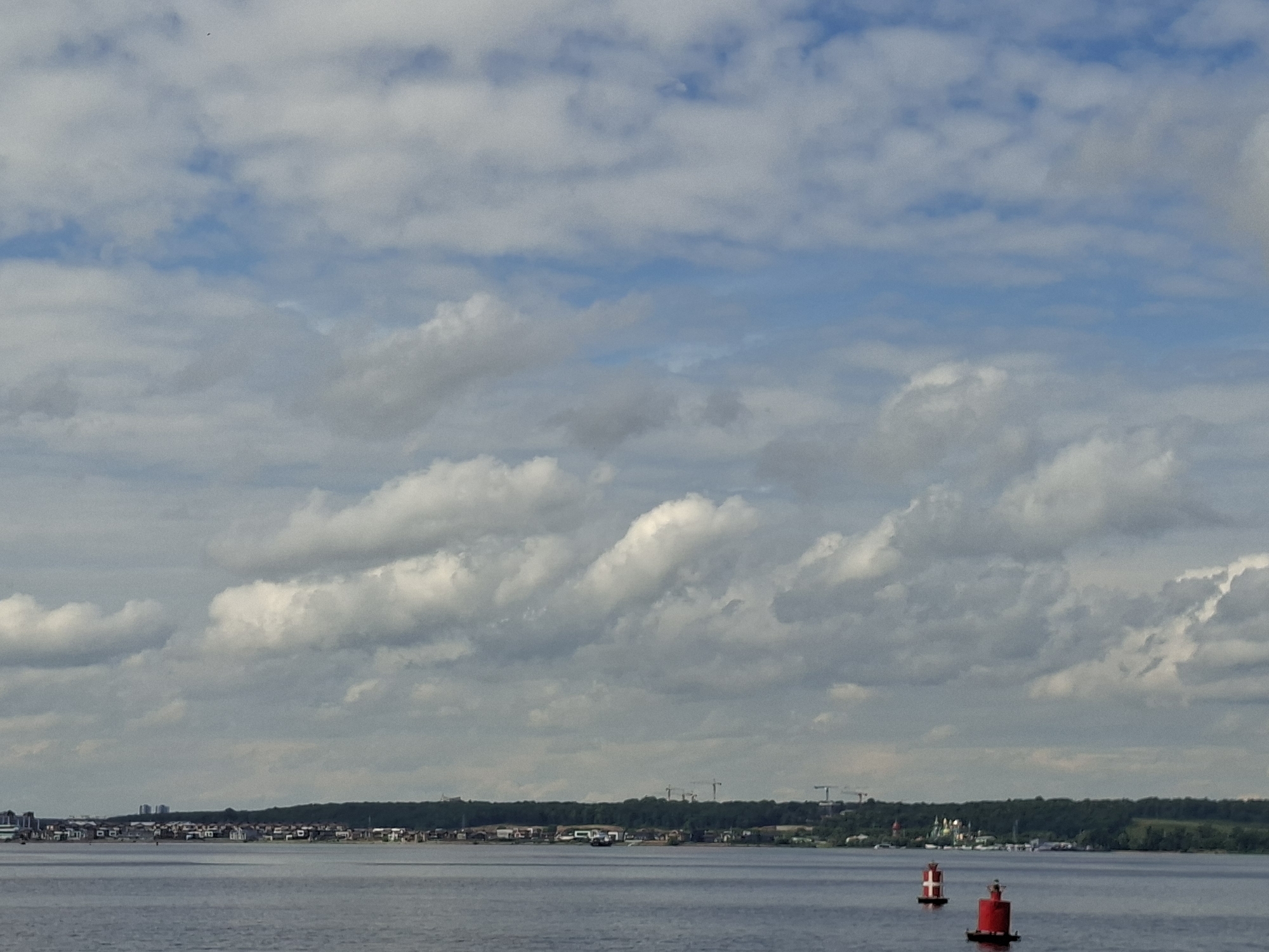 The image depicts a wide shot of a body of water, likely a river or lake, with a distant shoreline. The sky is partly cloudy with cumulus formations against a blue backdrop. On the shoreline, a strip of trees forms a dark green band, behind which various structures are visible, including buildings and what appear to be construction cranes. The foreground of the water features two buoys, one red with white markings and another primarily red. The overall scene suggests a moderately populated area with ongoing development, viewed from a distance across the water. The location is identified as Verhnij Uslon, Russia.